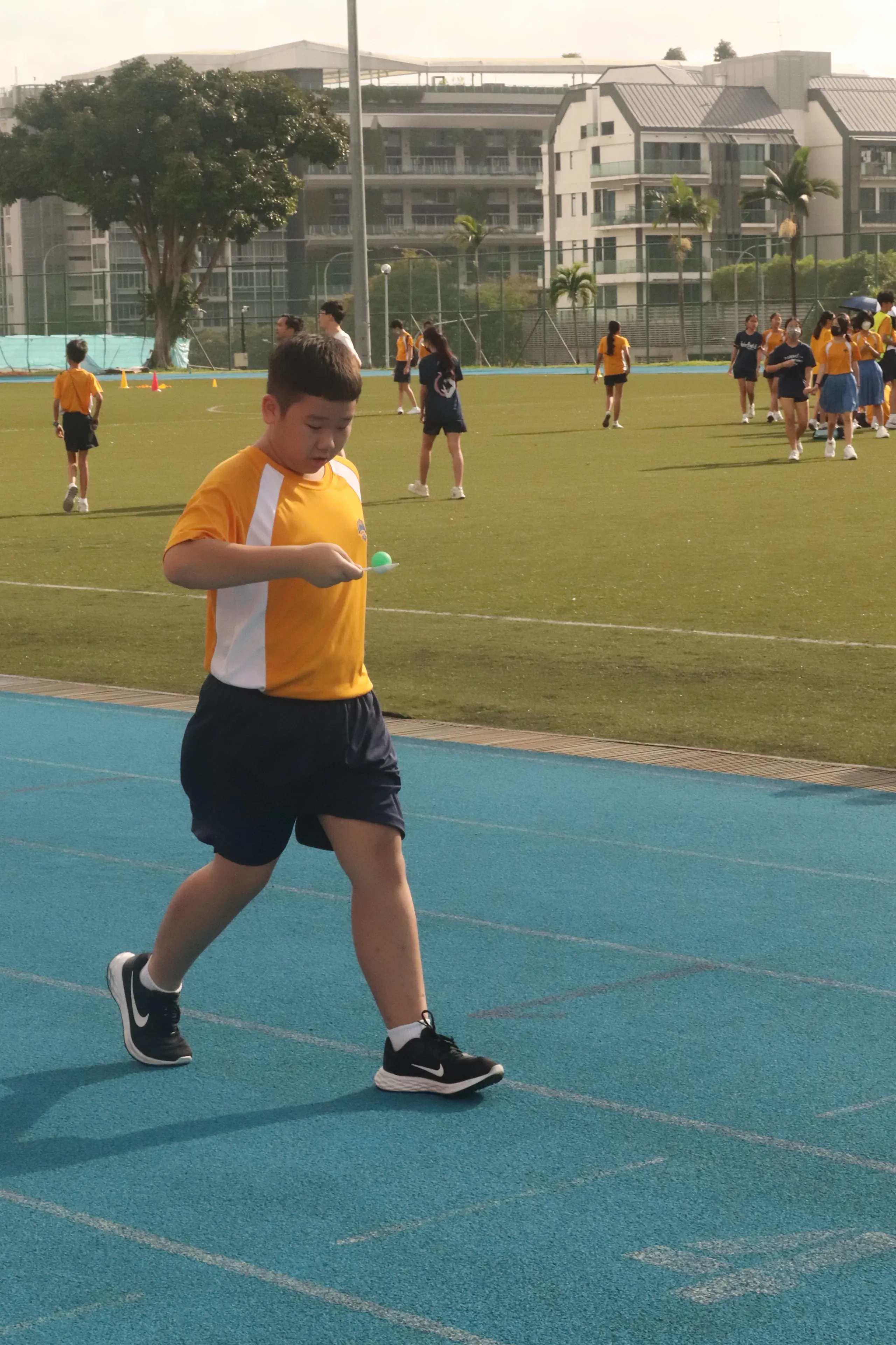 A male secondary school student in a yellow and white PE uniform carefully balancing a green ping pong ball on a white spoon while walking briskly down a blue running track. Other students and campus buildings at Ngee Ann Polytechnic are visible in the background under bright daylight.