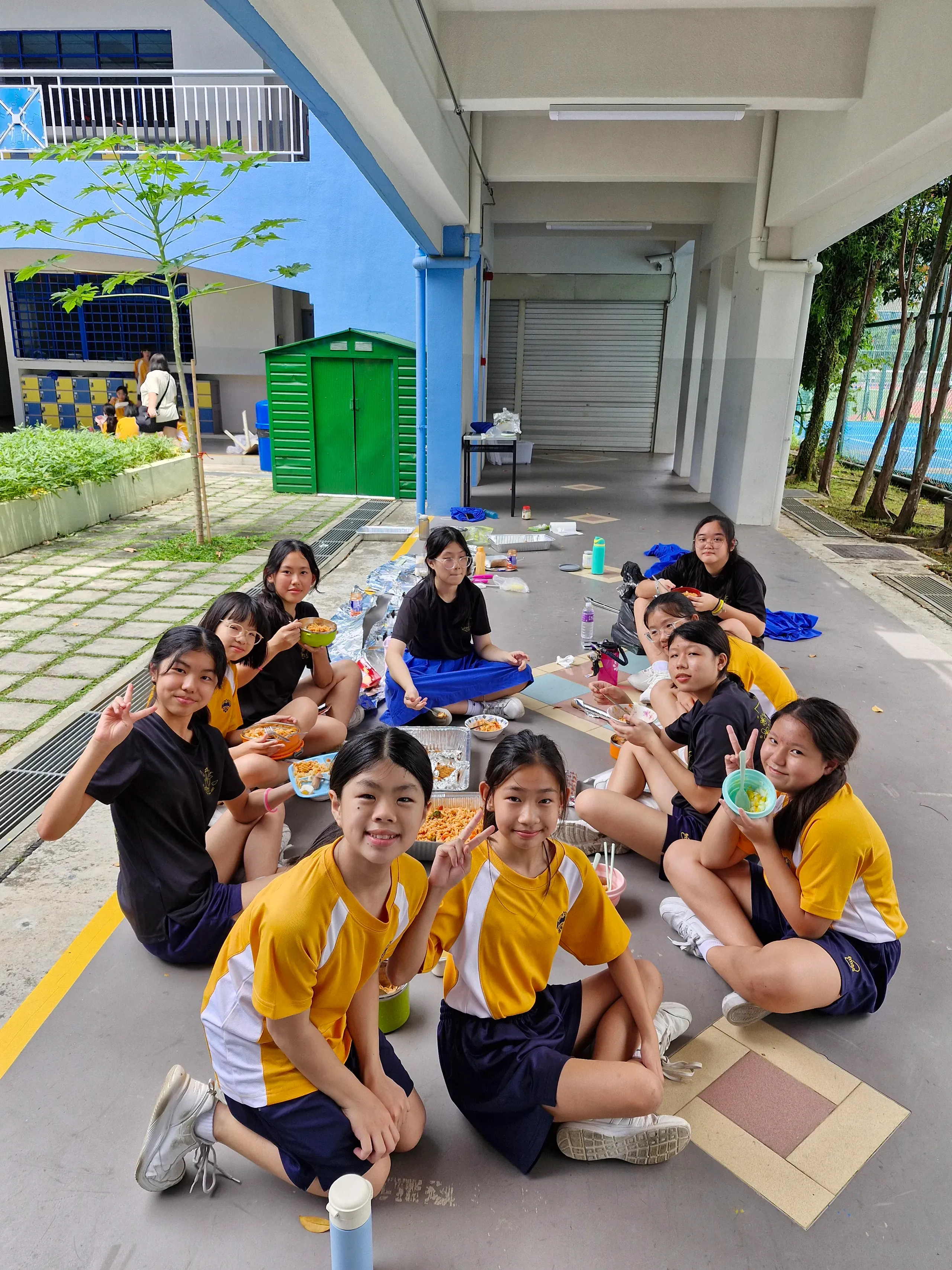 A group of female Fairfield students in yellow and white PE jerseys and black t-shirts sit in a circle on the floor of a sheltered school walkway, smiling and flashing peace signs while sharing a communal picnic meal from catering trays.