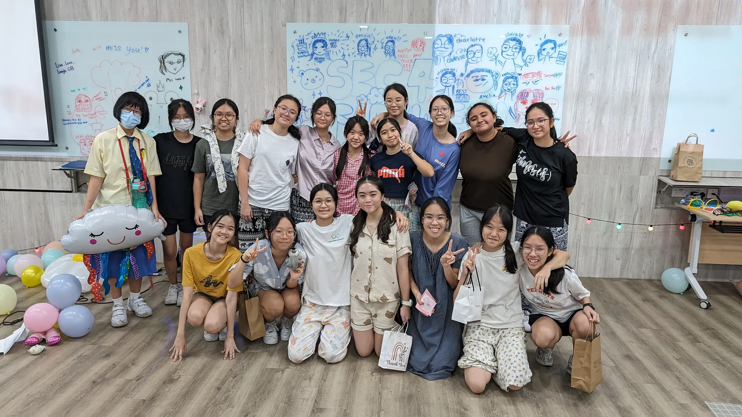 A large group of female Fairfield students in casual clothing and pajamas smile and pose together for a group photo indoors. They are gathered in front of a white glass whiteboard filled with drawings and text that reads "SEC 4." Colorful balloons and gift bags scatter around the room.
