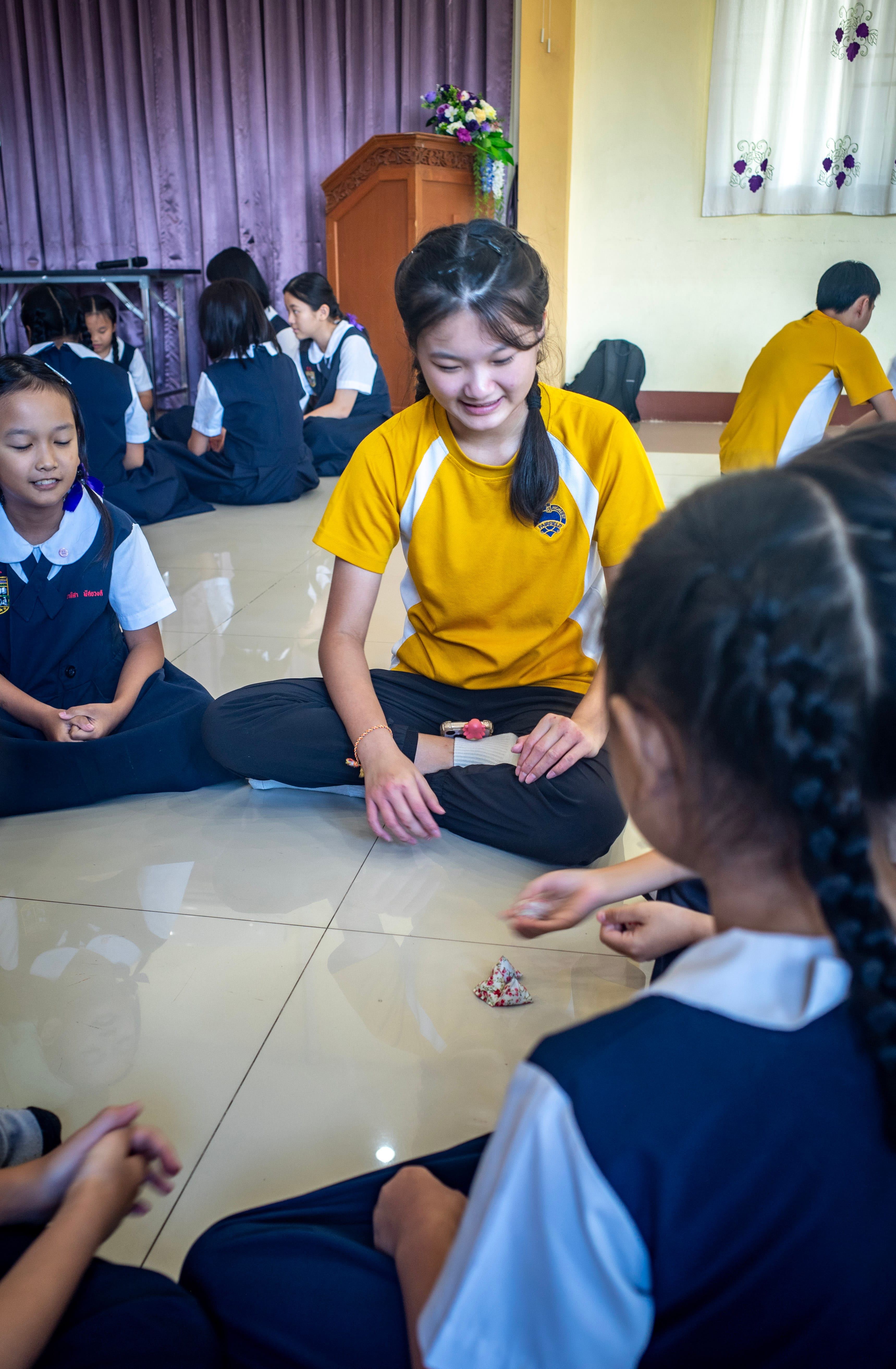A female Secondary 3 student wearing a yellow and white school sports t-shirt sits cross-legged on a polished tiled floor, smiling warmly as she interacts with local Thai primary school students. The primary school children are dressed in dark blue pinafores or uniforms over white collared shirts. They are gathered in a small circle playing a traditional tabletop game with small five-stone cloth beanbags. In the background, other groups of students are seated across the school hall near a wooden speaker's rostrum and light purple stage curtains.
