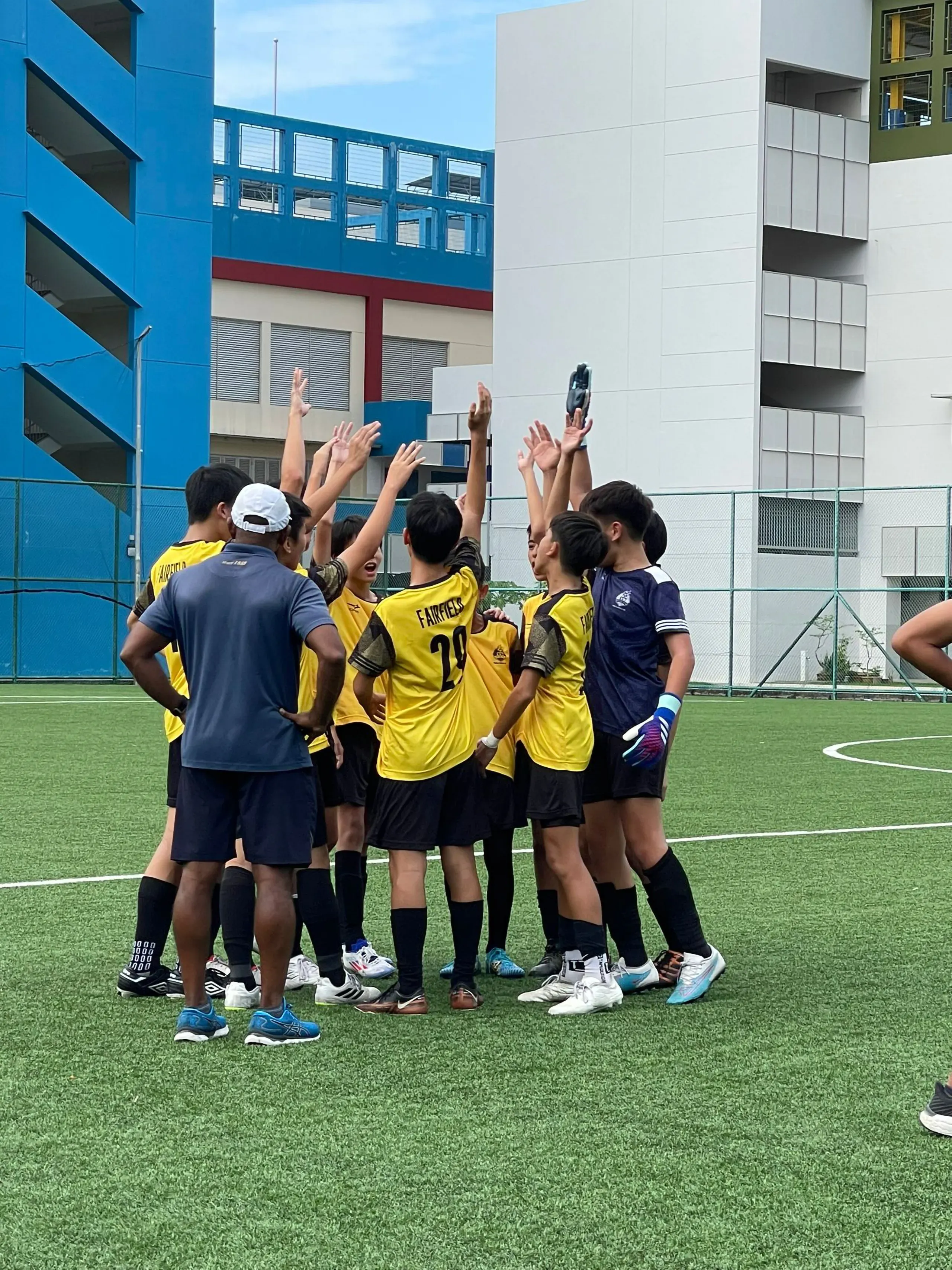 A group of Fairfield football players gather in a tight circle on an outdoor green turf field, raising their hands together in a unified team cheer. Most of the players are wearing yellow jerseys with black shorts and socks, including one showing the name "FAIRFIELD" and number 29 on the back. A goalkeeper in a dark blue jersey and colourful gloves stands on the right, while a teacher in charge, Mr Ravi in a dark blue polo shirt, black shorts, and a white cap watches from the foreground with his hands on his hips. Modern multi-story school buildings are visible in the background under a blue sky.