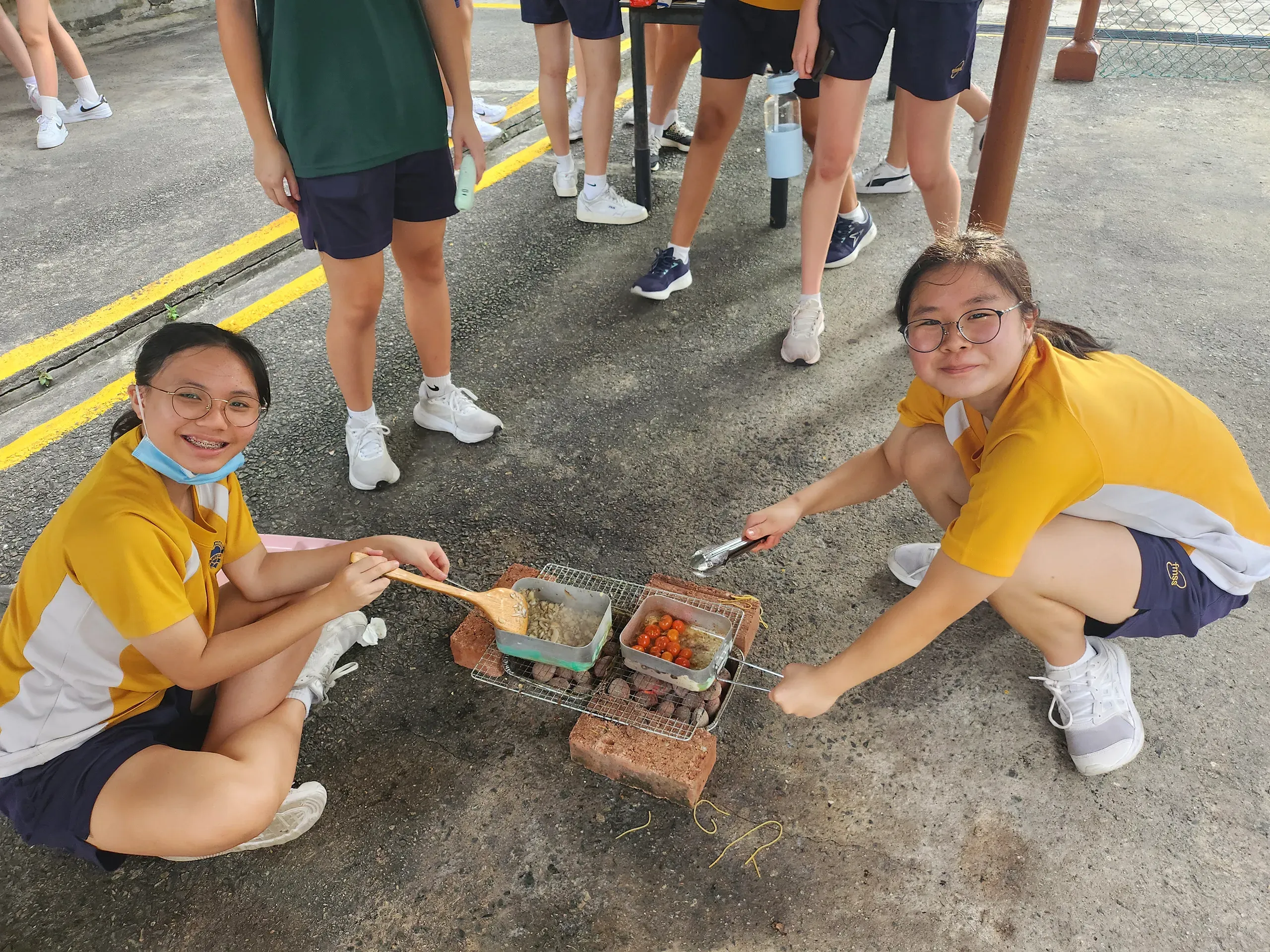 Two female student leaders smiling and looking up at the camera while outdoor cooking over an improvised charcoal grill during the training camp. Squatting on an asphalt ground, the student on the left wears glasses and a lowered medical mask, using a wooden spatula to stir food in a metal mess tin. The student on the right, also wearing glasses, uses tongs to manage cherry tomatoes grilling in a second mess tin. Both are dressed in yellow and white school sports shirts and dark shorts. In the blurred background, other camp participants in sports attire stand around and chat.