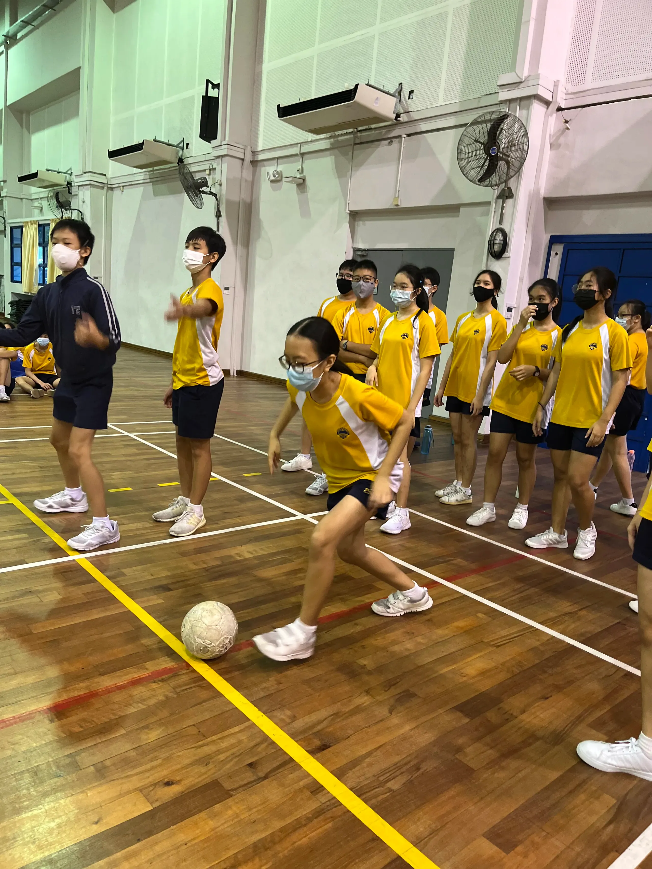Secondary 2 students wearing face masks and yellow-and-white sports t-shirts participate in a physical activity on the wooden floor of a school hall during FLEX week. In the foreground, a female student with glasses steps forward to control a football, while her classmates line up on the side watching and waiting for their turn.