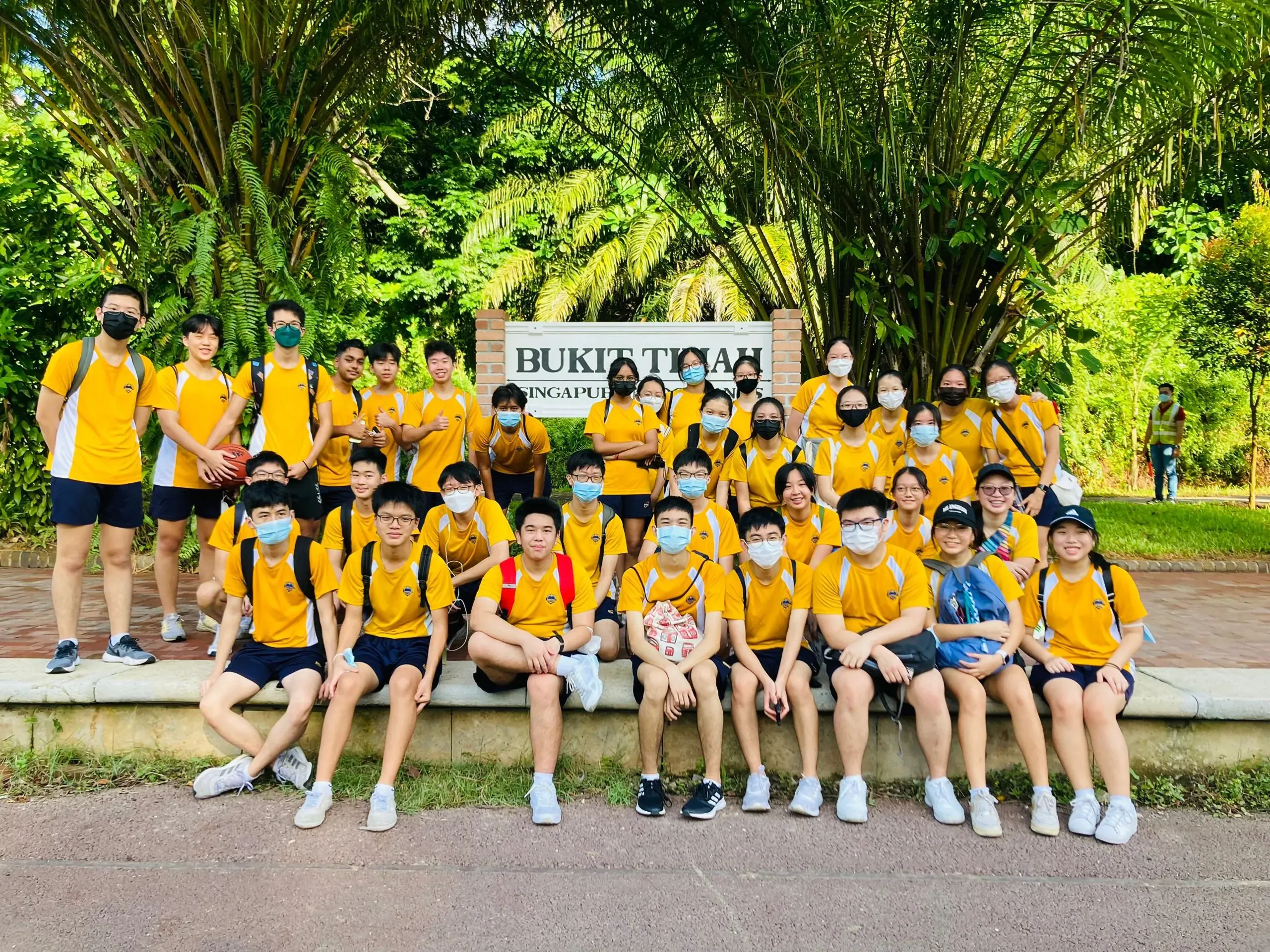 A large group photo of secondary school students posing together outdoors in front of a white brick sign that reads "BUKIT TIMAH". The students are wearing yellow and white school PE uniforms and protective face masks. A few students stand on the sides, while the majority sit in a line along a low concrete ledge with dense green trees and tropical foliage filling the background under bright daylight.