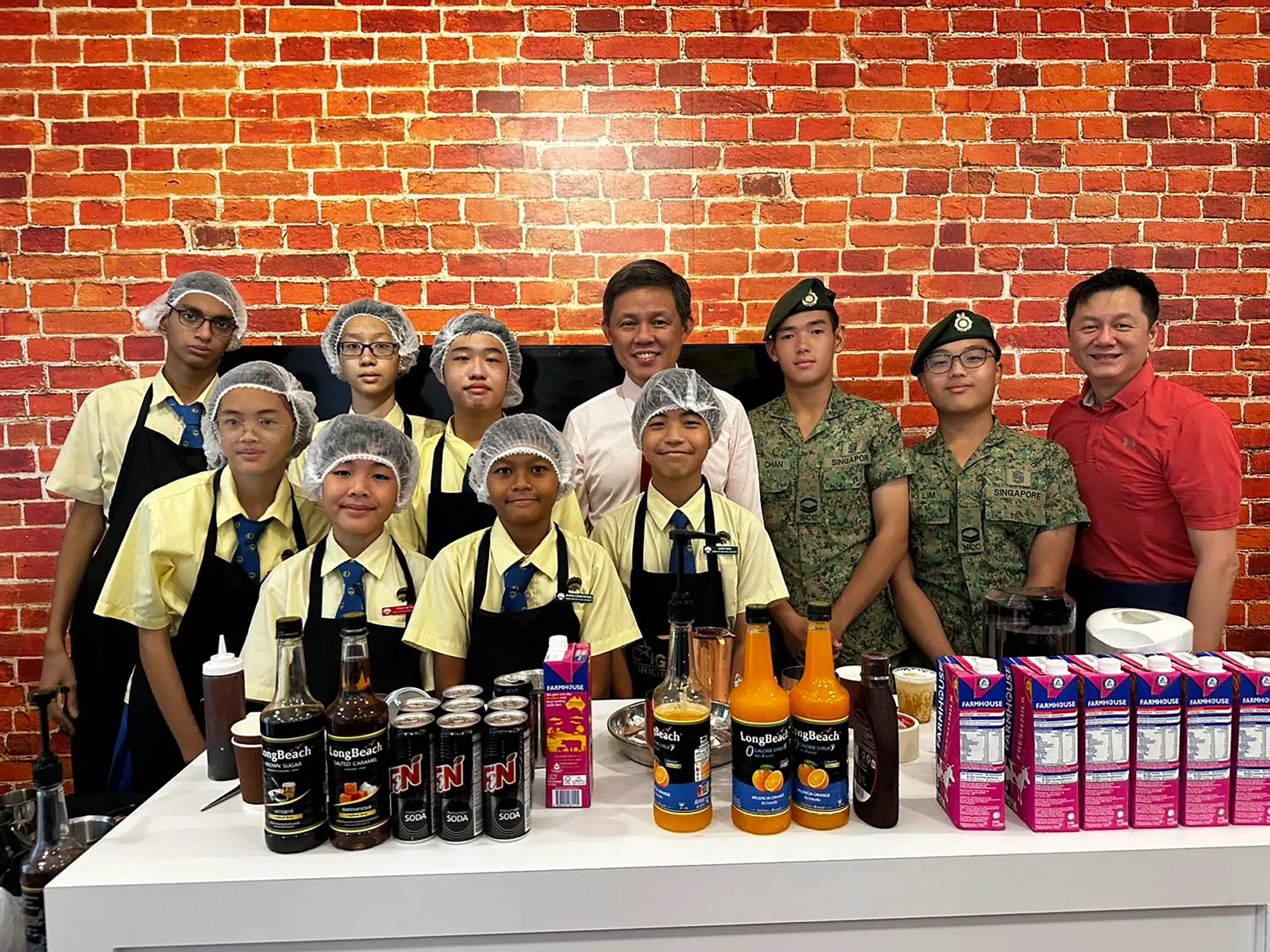 Minister for Education Mr. Chan Chun Sing stands in the center of a group portrait behind a mocktail serving counter against a rustic red brick wall. He is surrounded by seven Fairfield student mocktail baristas wearing yellow uniforms, black aprons, and hairnets, alongside two NCC land cadets in green camouflage uniforms and berets. On the far right, a supportive parent in a red polo shirt smiles for the camera. The counter in the foreground is neatly lined with beverage cartons, soda cans, and flavor syrups.