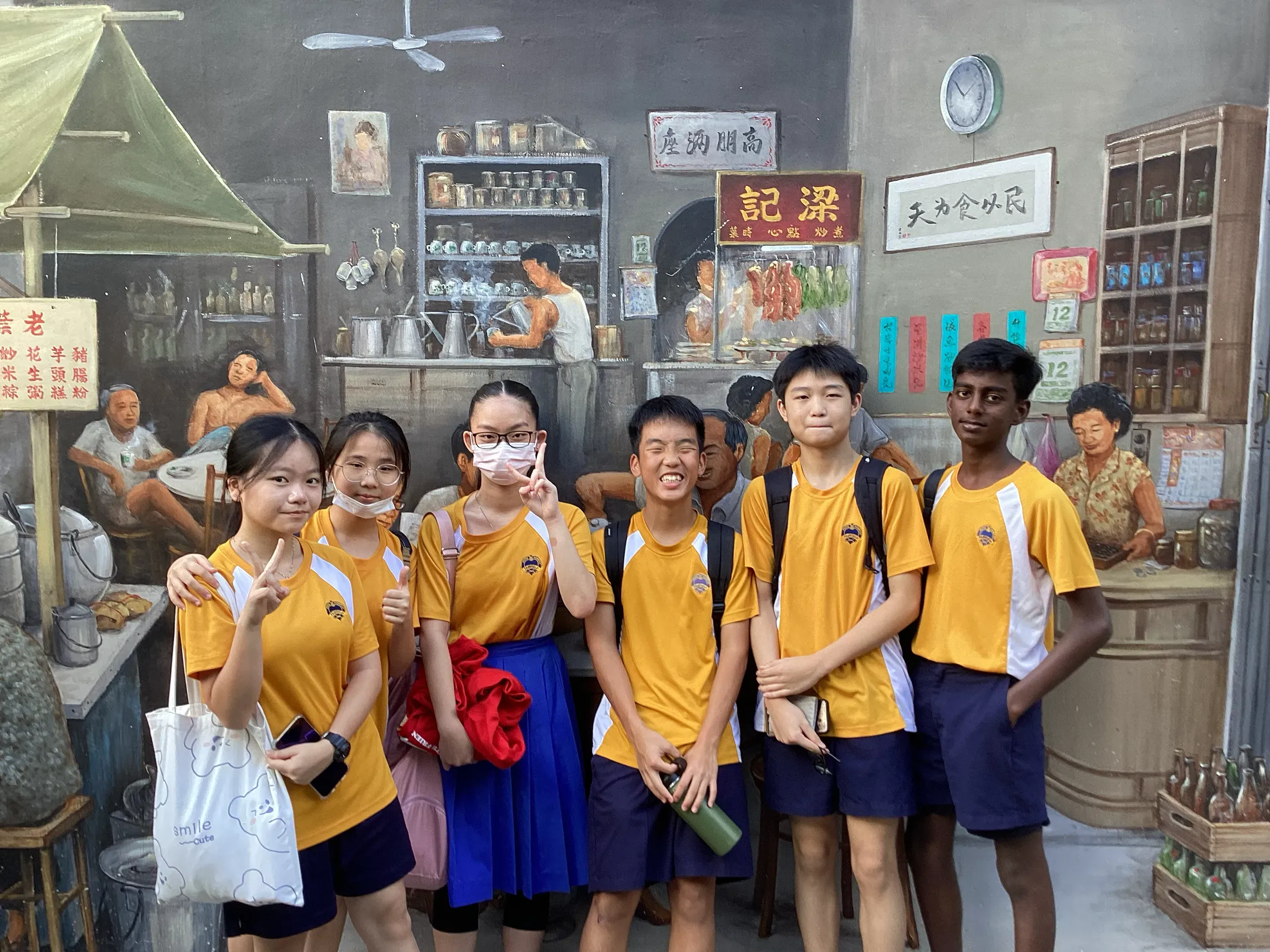 Six Secondary 2 students wearing yellow and white school PE shirts posing together in front of a detailed, life-sized heritage wall mural. The artwork depicts a traditional old Singaporean coffee shop (kopitiam) and roadside hawker stalls, with characters from the past painted in the background.