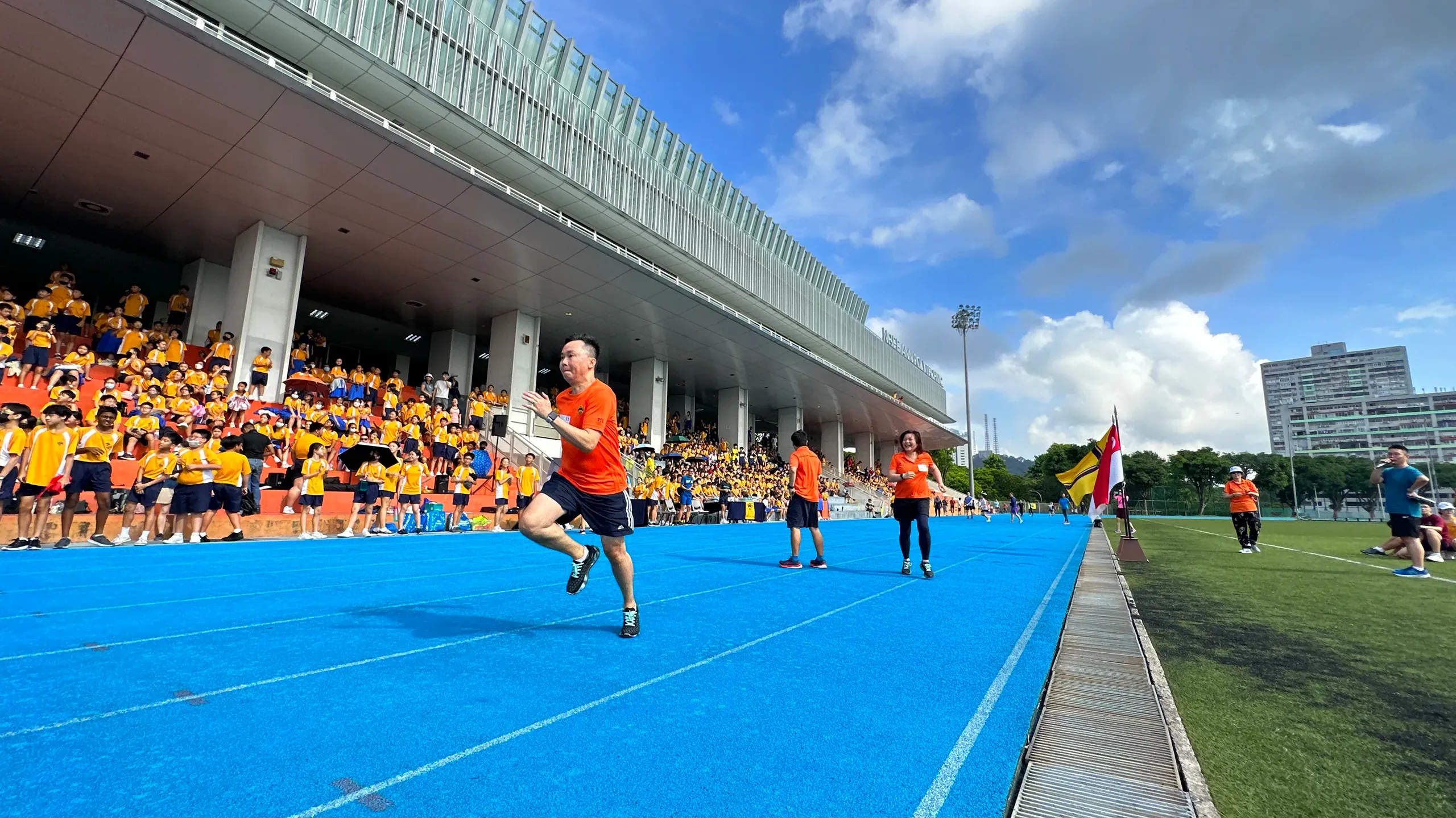 A wide, low-angle shot of a parent race taking place under a blue, cloudy sky on the blue running track at Ngee Ann Polytechnic. A father in an orange athletic t-shirt sprints hard down the track lane in the foreground, while a mother in an orange polo shirt runs behind him. A large crowd of students fills the grandstand to watch the race.