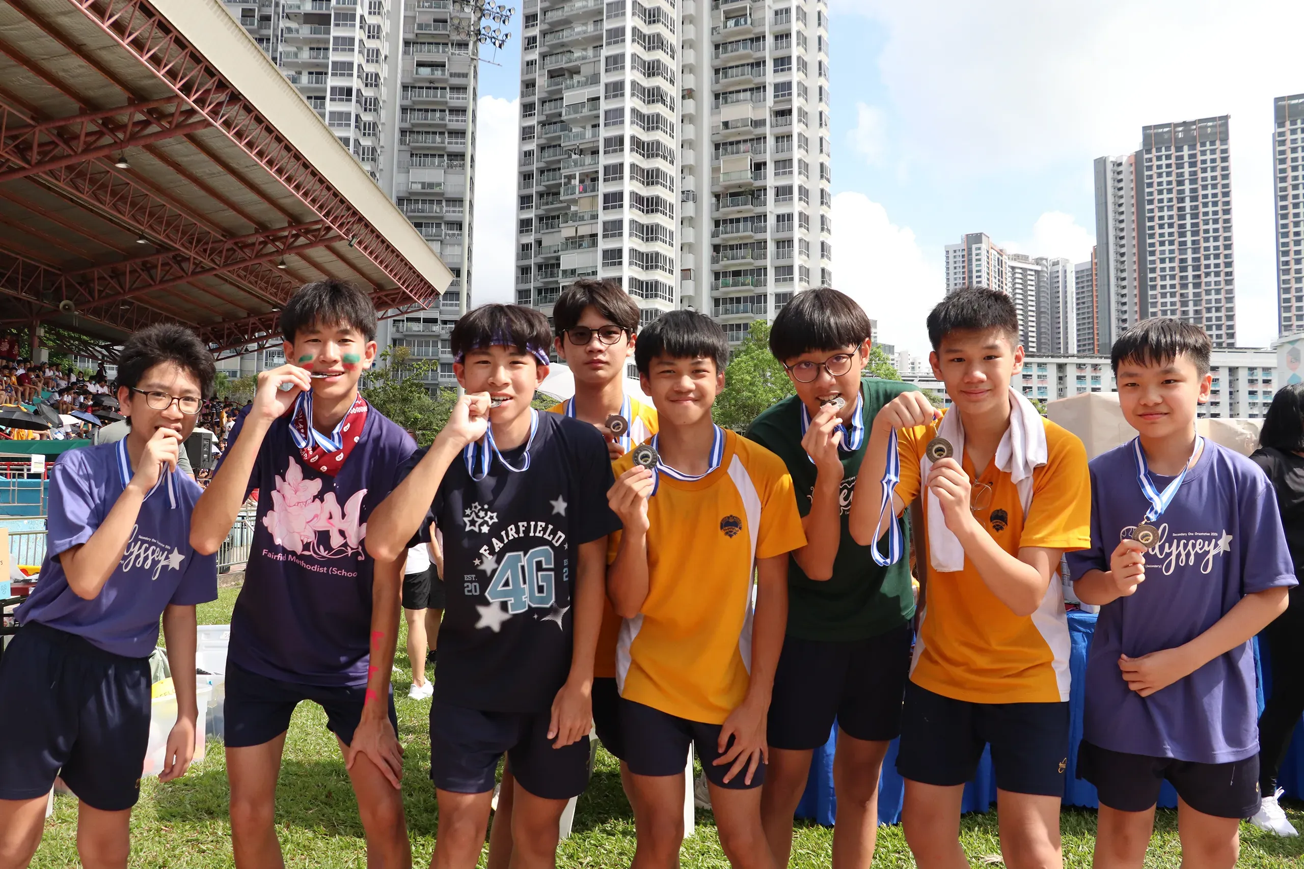 A group of eight male students stand shoulder-to-shoulder on a grass field posing for a group photo during Fairfield Methodist School (Secondary) Sports Day 2026. Smiling proudly, they hold up and bite their newly won sports medals. The students are wearing various colored house t-shirts, with stadium grandstand seating and tall residential apartment buildings visible in the background under a blue sky.