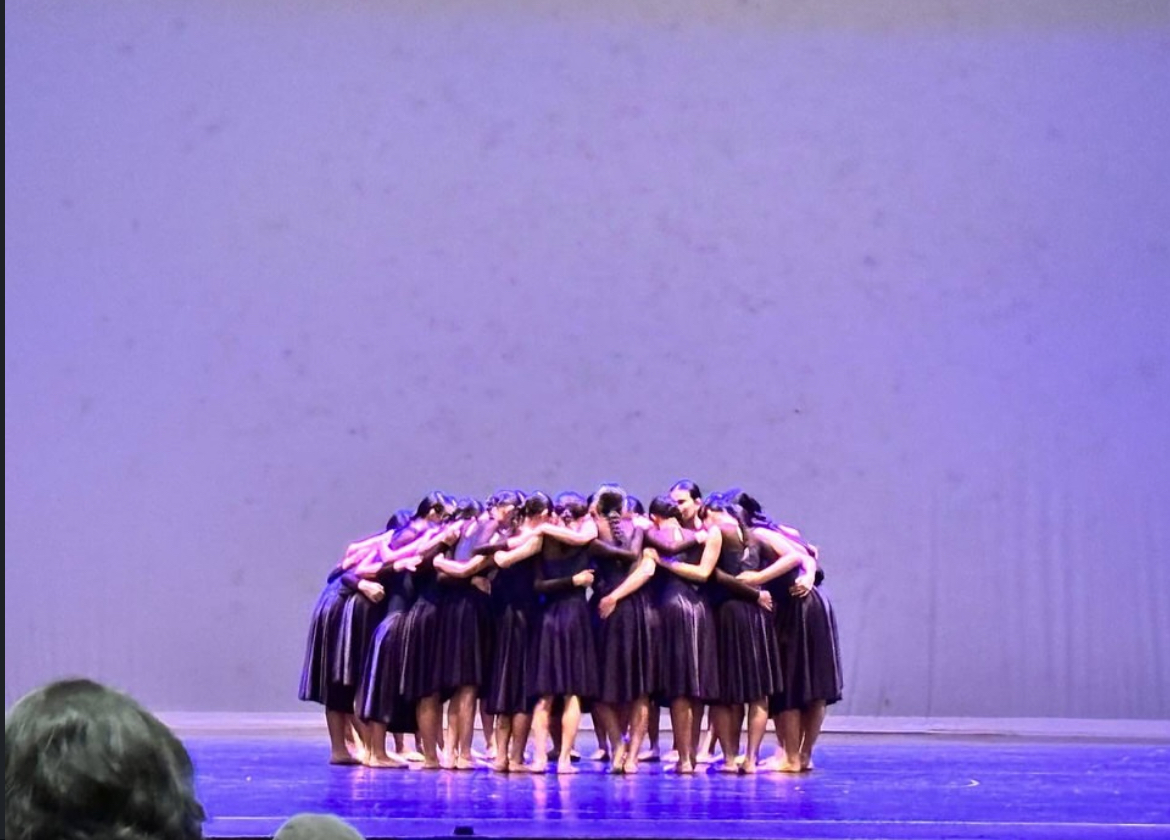 A close group shot of the school dance troupe huddled tightly in a circle backstage before their SYF performance. The dancers are wearing white long-sleeved costumes with flowing blue sashes and have their hair neatly styled in low buns. They are smiling warmly at each other with their arms wrapped around one another's shoulders in a supportive huddle, with several hands placed together in the center. The background shows a dimly lit backstage hallway area.