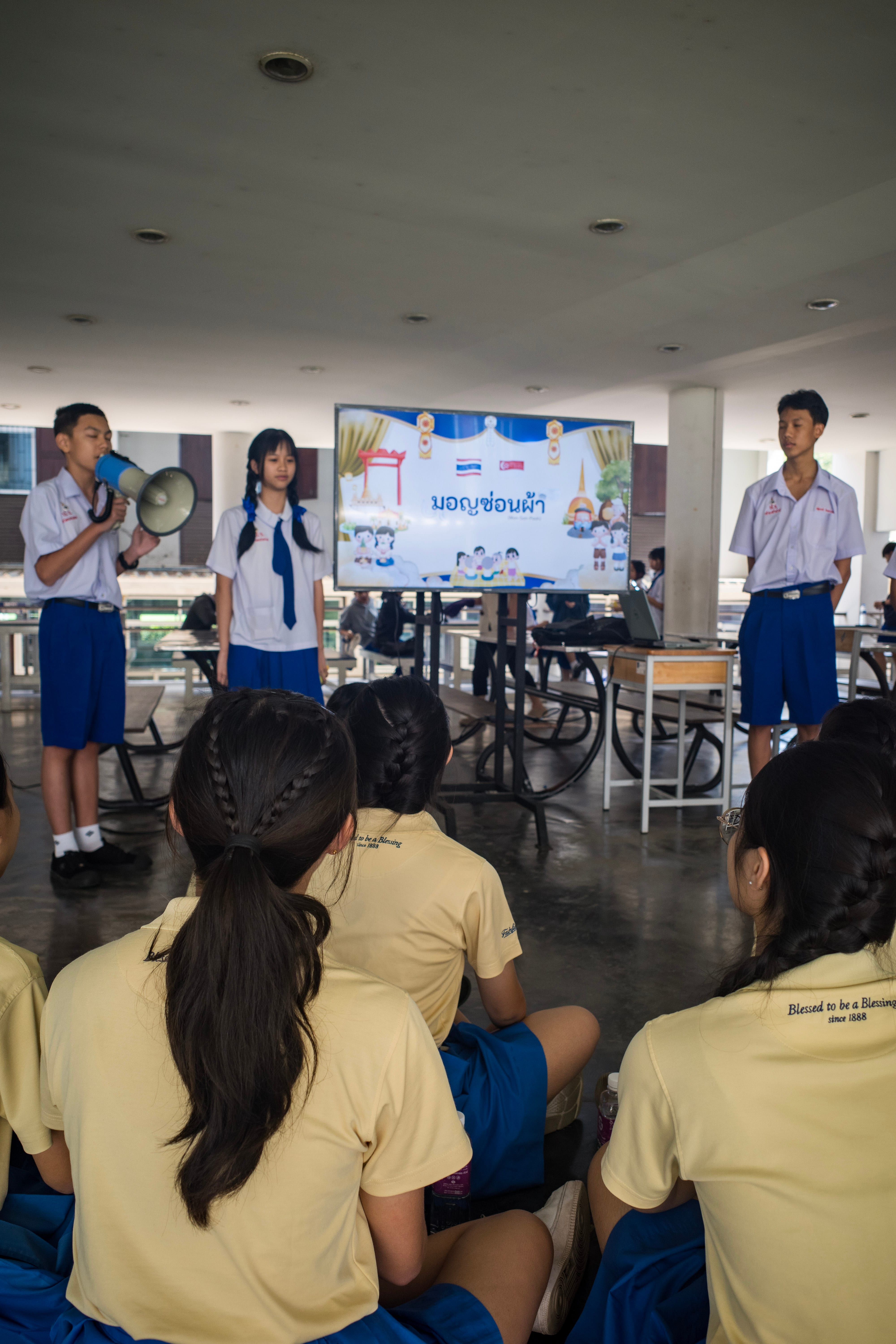 A group of Secondary 3 students in yellow school shirts and blue skirts sit on the floor from a back-facing view, listening to a presentation by local Thai students during their TIE trip. In the background, a Thai male student stands on the left speaking into a blue-and-white megaphone, while a female student and another male student in traditional white and blue Thai school uniforms stand at attention nearby. Between them, a large television screen on a rolling stand displays a colorful presentation slide featuring illustrations, the Singapore and Thailand flags side by side, and Thai script text.