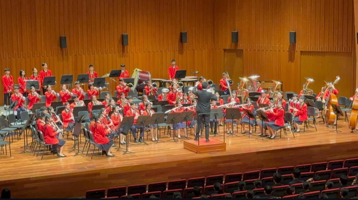 A wide-angle view of the school symphonic band performing on stage during the Singapore Youth Festival (SYF) inside a grand, wood-paneled auditorium. The student musicians are seated in semi-circular rows, dressed in vibrant formal red blazers, paired with blue skirts or dark trousers. Woodwind sections are positioned at the front, brass players are in the center, and percussionists stand at the rear near a large bass drum. In the middle foreground, a conductor dressed in a black formal suit stands on an elevated wooden podium with his back to the empty, red-upholstered theater seats, actively directing the performance.