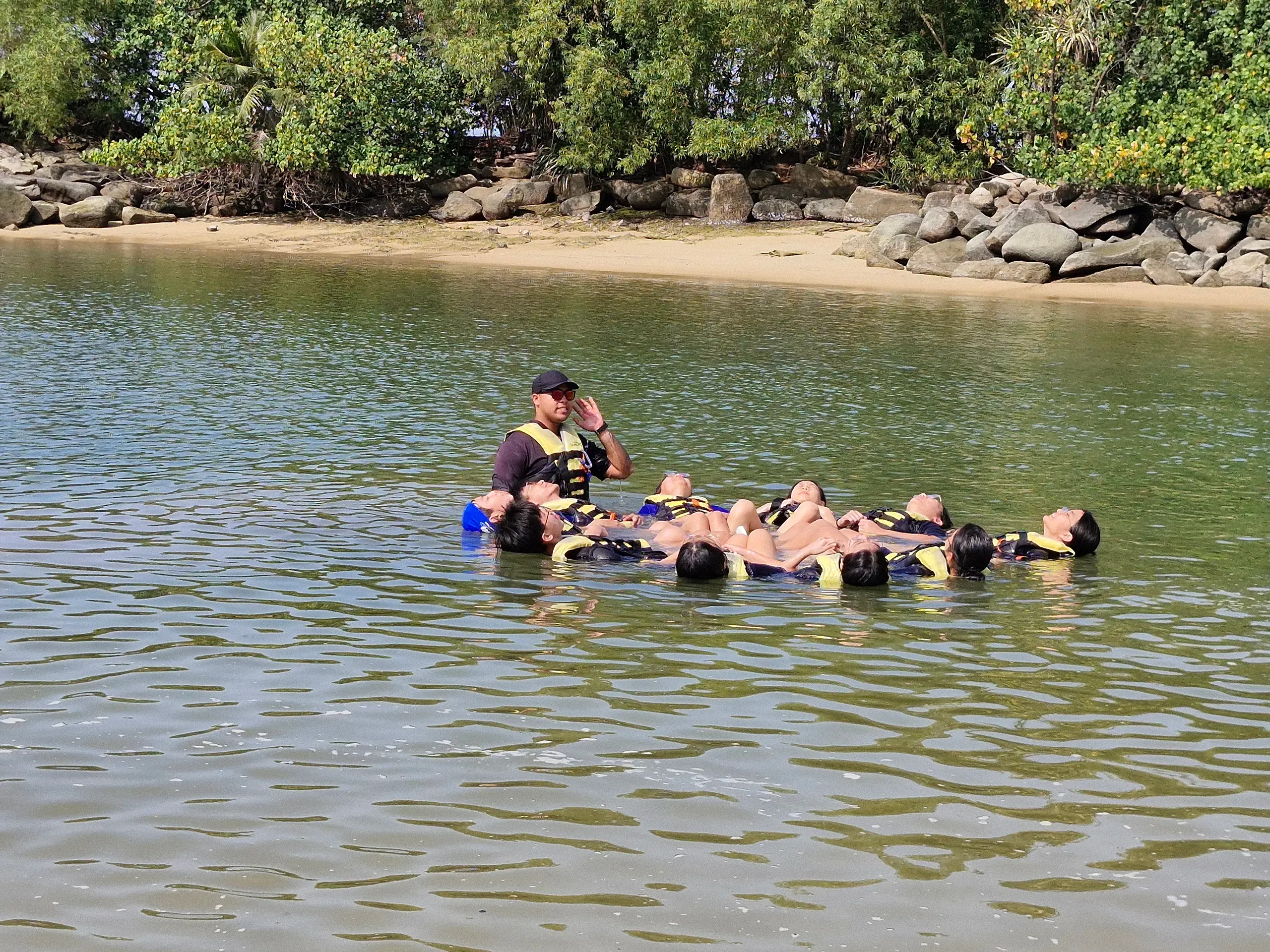 An outdoor camp instructor wearing a black cap, sunglasses, and a life jacket stands in a shallow coastal waterway, guiding a group of ten student leaders during a water safety activity. The student leaders are floating calmly on their backs in a tight, connected circular formation while wearing yellow and black "SeaOPS" life jackets. In the background, a serene sandy shoreline is lined with large rocks and thick tropical green trees under a bright, sunny sky.