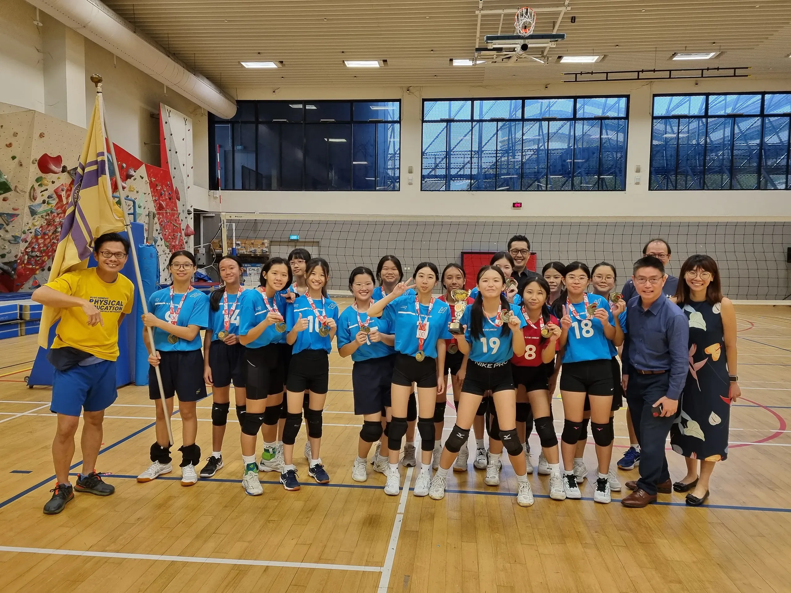 The Fairfield girls' volleyball team poses for a celebratory group photo on an indoor sports court alongside their coaches and teachers and school leaders. The players wear blue athletic jerseys, black shorts, and medals around their necks, with one player in the center proudly holding a championship trophy. On the far left, our HOD PE, Mr Francis Ng in a yellow shirt holds up a large school flag, while a climbing wall and volleyball net are visible in the background.