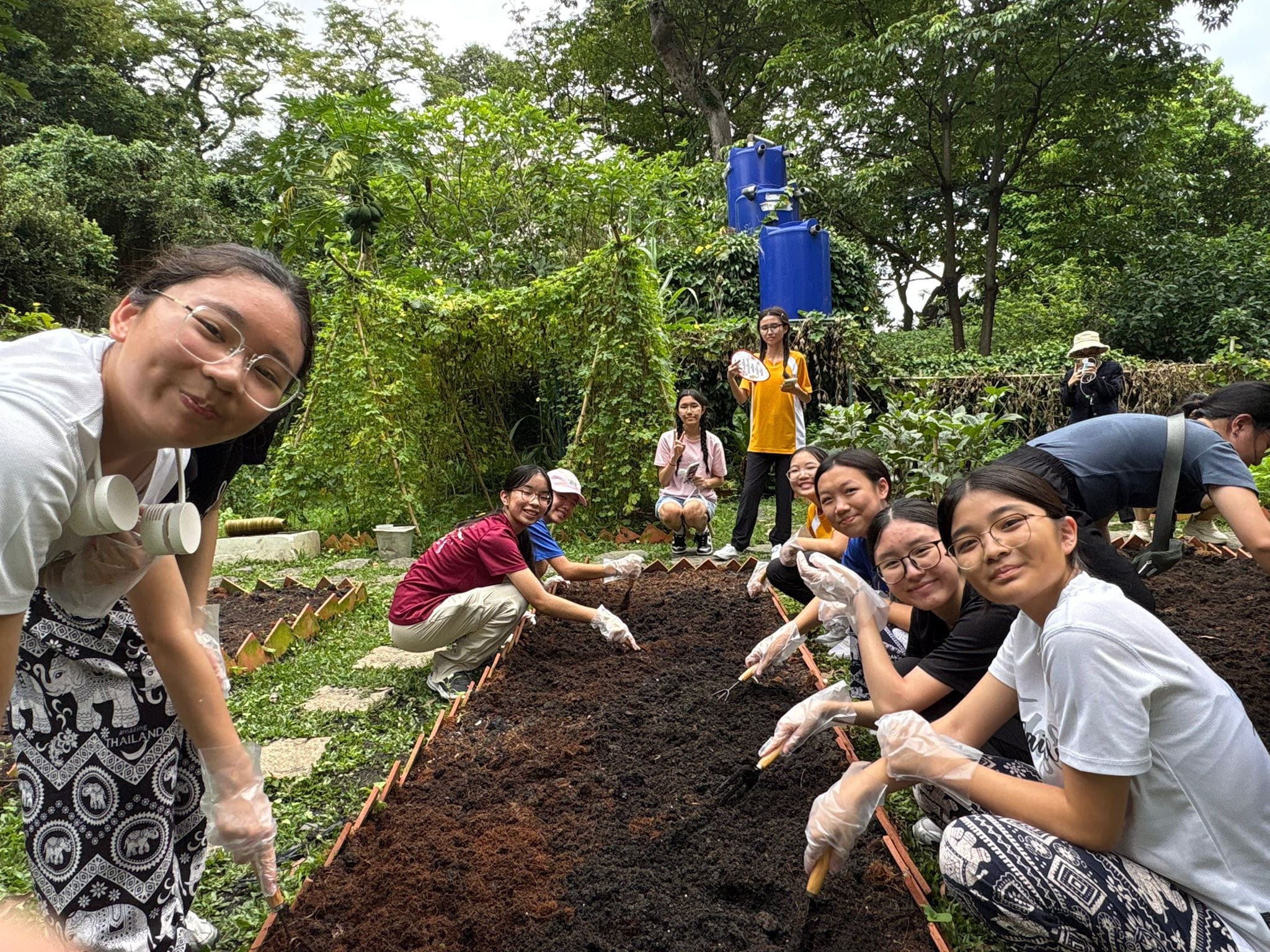 group of Secondary 3 students participating in an outdoor agricultural activity at an eco-farm or garden during their TIE trip to Thailand. Several female students wearing casual shirts and plastic gloves kneel along both sides of a long, freshly tilled soil plot, using small hand tools to tend the earth. A student on the left, wearing traditional black-and-white patterned Thai elephant pants and a white portable neck fan, smiles closely at the camera. In the background, lush green trees, garden trellises, two large blue water storage tanks, and other classmates observing the session fill the vibrant outdoor space.