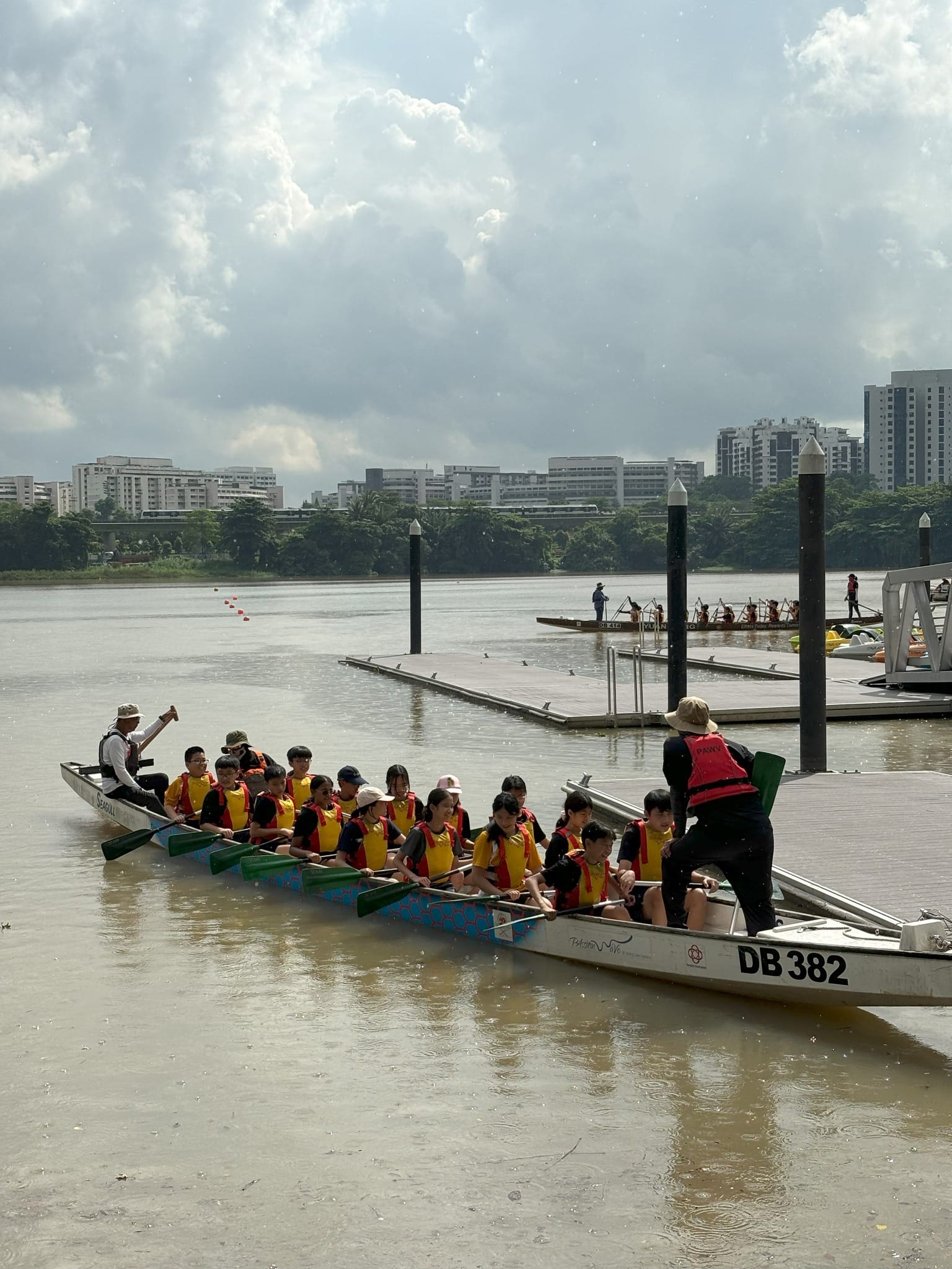 A group of Secondary One students sit in a long, white dragon boat labeled "DB 382" on the water near a floating jetty during their school camp. The students wear yellow school sports shirts and bright yellow life jackets while holding paddles across the boat's rim. A camp instructor wearing a red life jacket and a sun hat stands at the bow facing them, while another instructor coordinates from the stern. The background features a wide river or reservoir, floating dock platforms, a tree-lined bank, and distant high-rise residential buildings under a bright, cloudy sky.