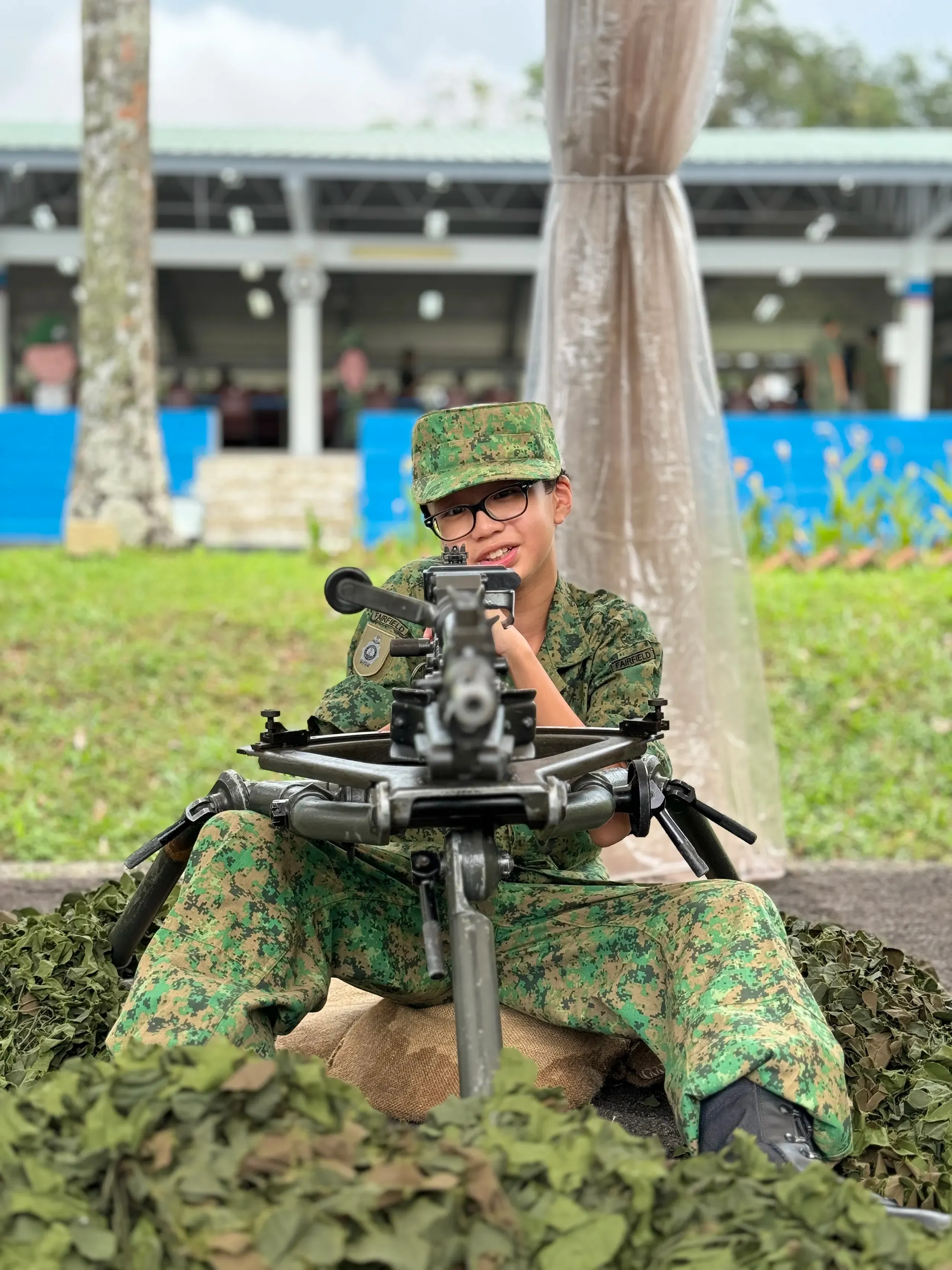 A young Fairfield NCC cadet wearing a green pixelated camouflage uniform, cap, and glasses sits behind a heavy machine gun replica mounted on a tripod behind a simulated camouflage net structure outdoors, looking forward intently.