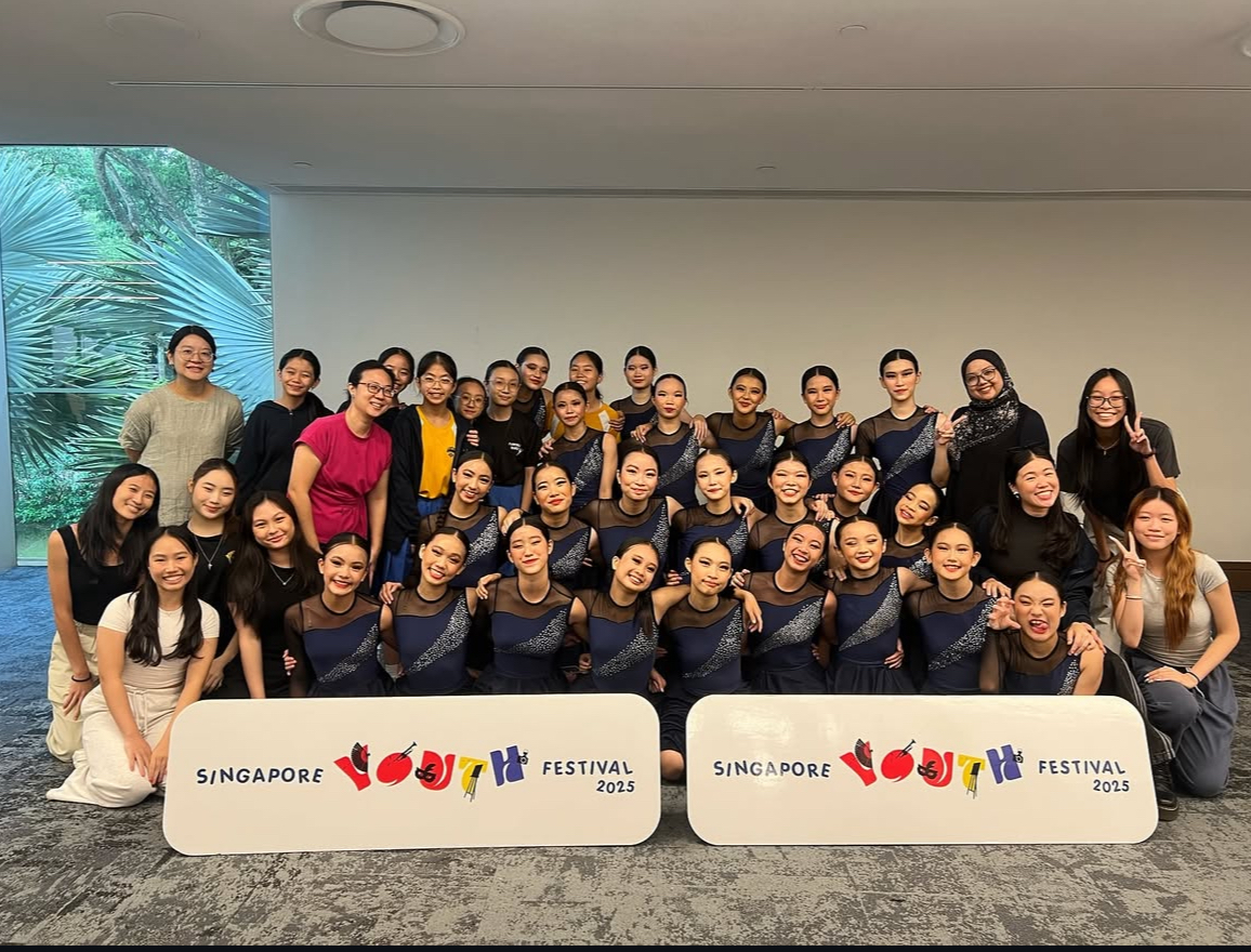 A group commemorative photo of the school dance troupe posing indoors after their performance for the Singapore Youth Festival (SYF) 2025. The dancers are kneeling and standing in rows, wearing matching sleeveless dark navy performance dresses with a diagonal, glittering silver-and-blue sequined sash design across the bodice. Their hair is tied back uniformly, and they wear stage makeup. In the front row, two matching white "Singapore Youth Festival 2025" signs are displayed on the carpeted floor. Teachers and instructors in casual attire smile and pose on both the left and right flanks of the student group.