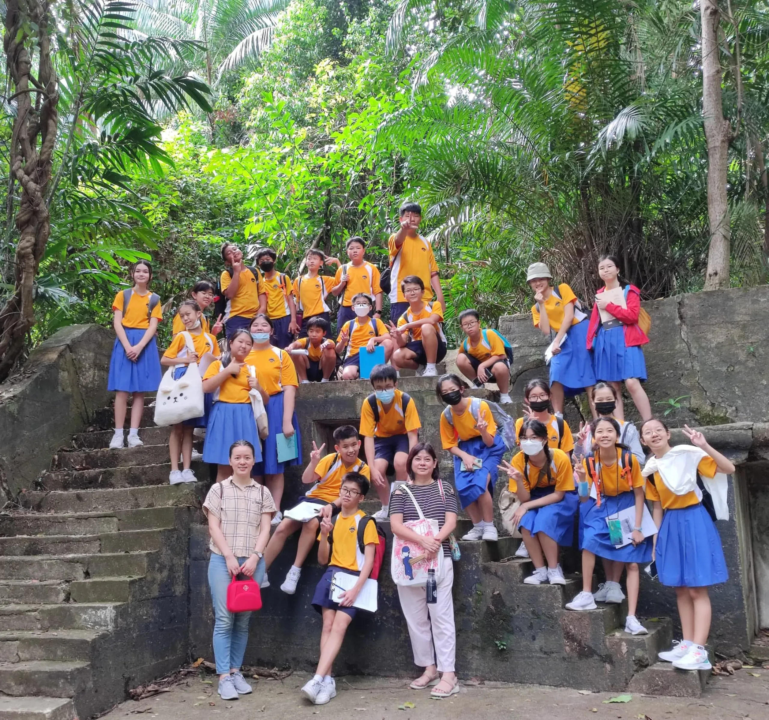 A large group of secondary 1 students and two teachers posing on concrete steps in a dense, green forested area during a learning journey. The students are wearing yellow and white school PE shirts paired with blue skirts or shorts, some carrying clipboards and backpacks. They are surrounded by lush tropical trees and vines.