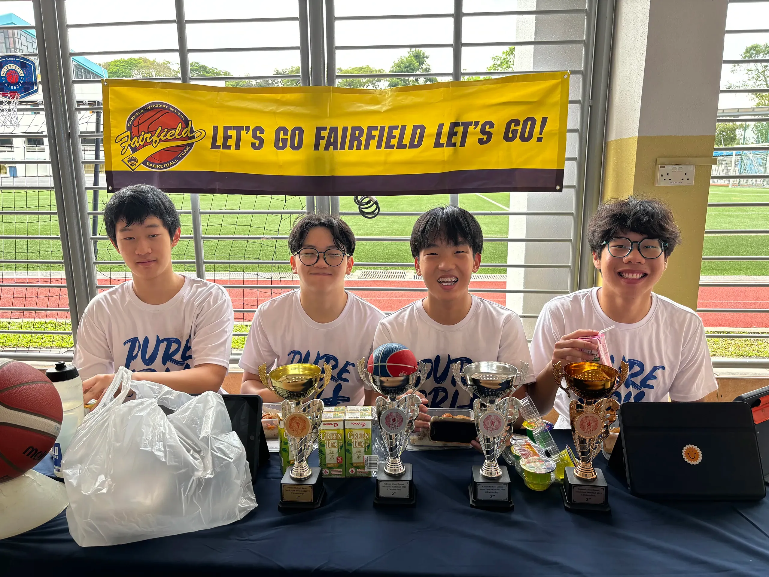 Four male Fairfield students in matching white t-shirts sit behind a table displaying sports trophies and refreshments. Hanging on the metal window gate directly behind them is a bright yellow banner that reads "LET'S GO FAIRFIELD LET'S GO!" with a basketball logo. On the table, four silver and gold trophies are lined up alongside drinks, snacks, and a tablet, with an outdoor running track and sports field visible in the background.