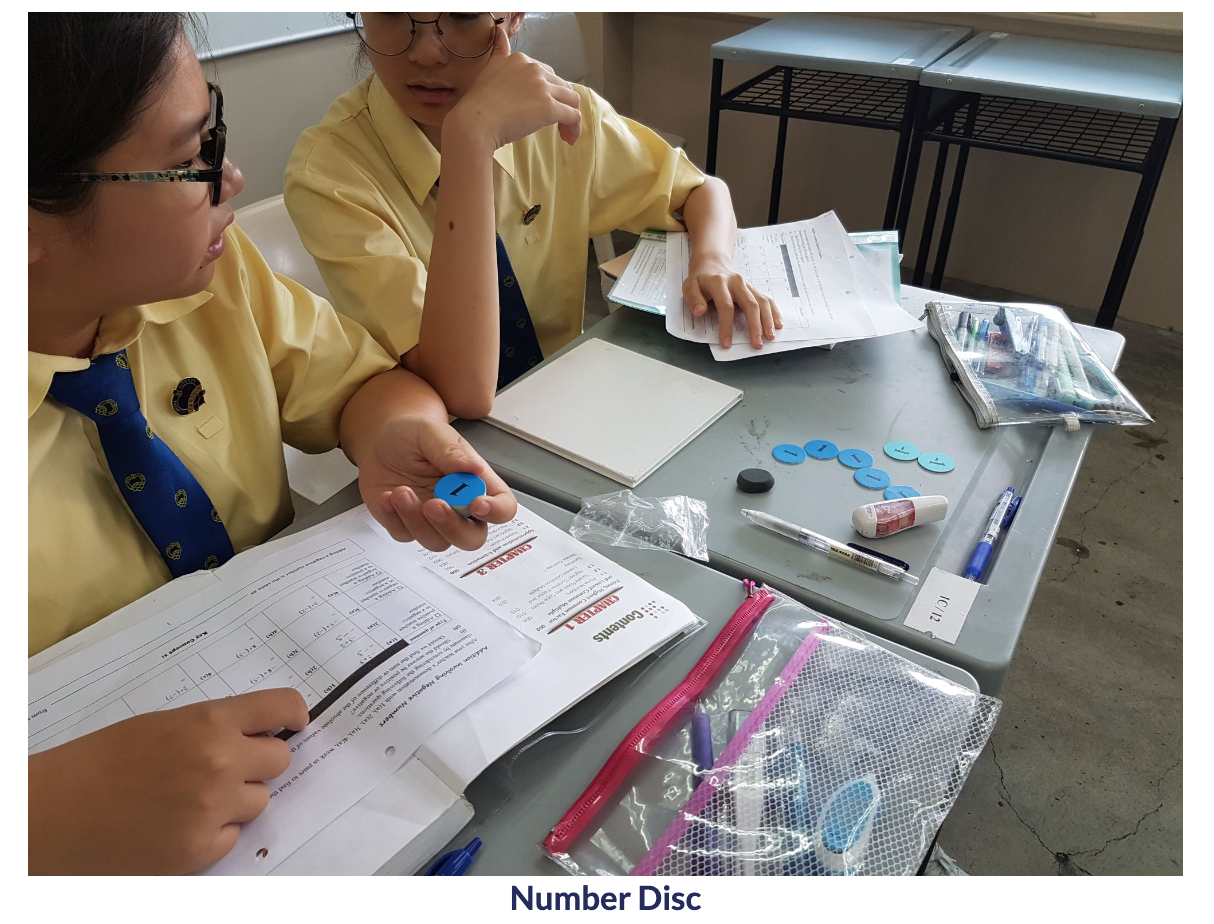 Two students in yellow uniforms sit at a desk using small blue number discs alongside open workbooks and pencil cases.