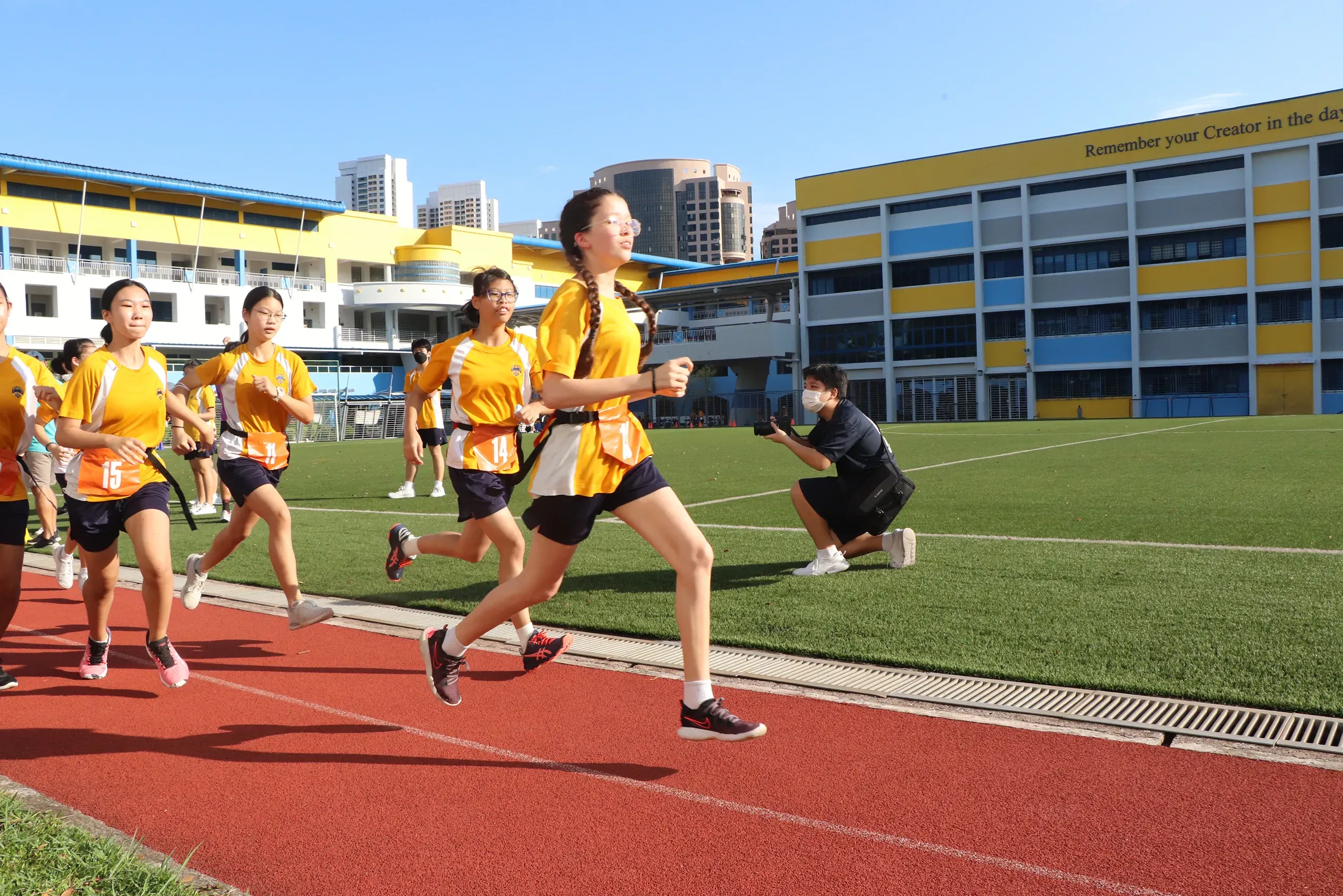 Female students wearing yellow sports shirts, dark shorts, and number tags running along the red athletic track during the Fairfield Methodist School (Secondary) Annual Run. A student photographer kneels on the artificial turf field in the background to capture the race, with the yellow and blue school buildings under a clear blue sky.