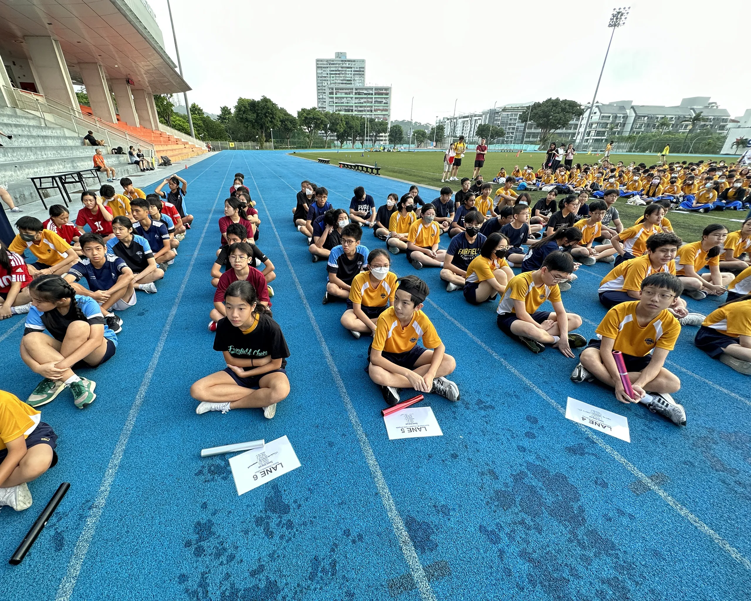Multiple teams of secondary school students seated in rows on a blue running track at the Ngee Ann Polytechnic sports complex, preparing for an Inter-CCA relay race. The front runners hold colored batons next to lane assignment papers labeled "LANE 4", "LANE 5", and "LANE 6", while a large crowd of student spectators watches from the grassy field in the background.