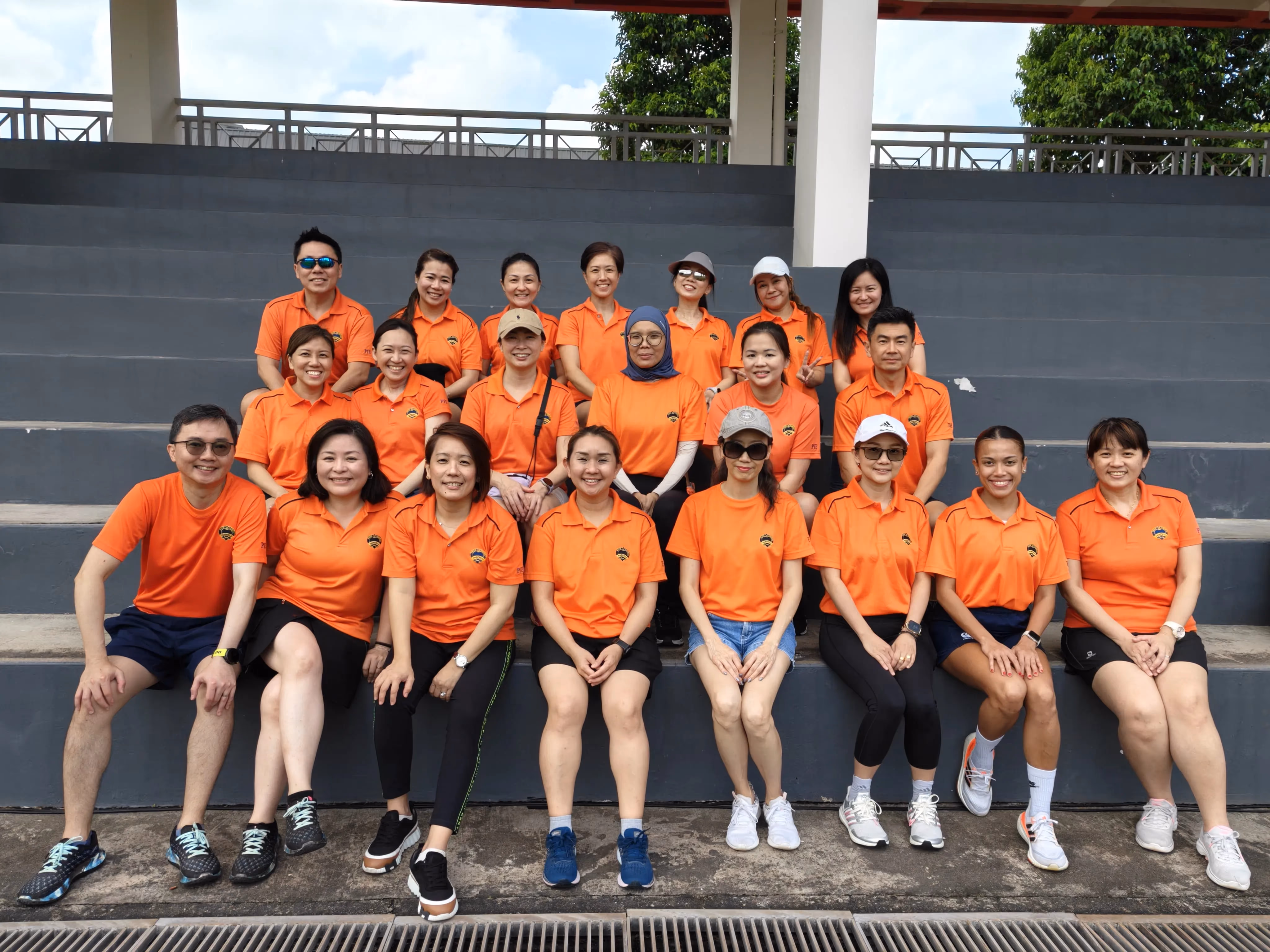 A group of about 20 people in matching orange polo shirts pose on stadium bleachers outdoors.