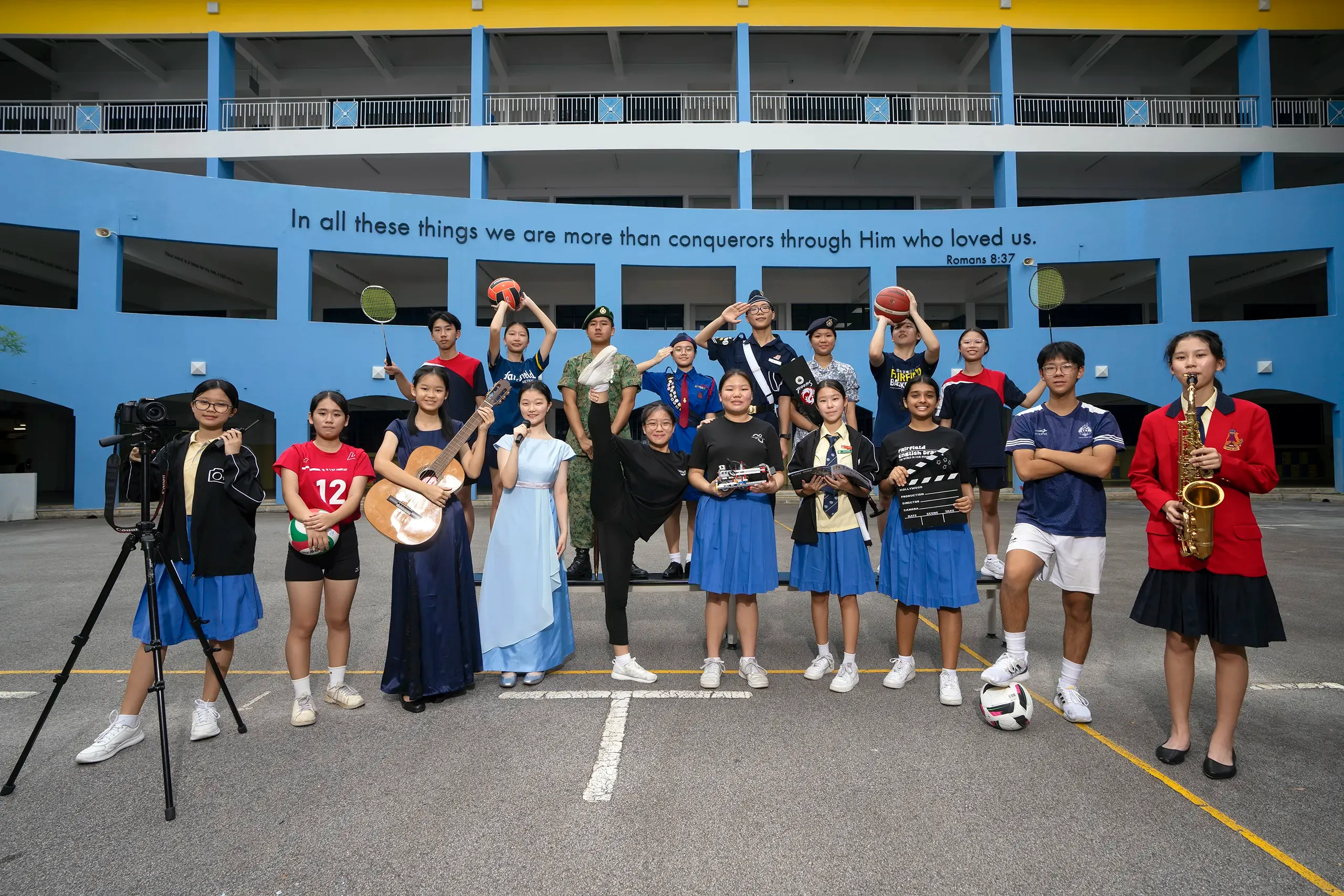 A large group of students in various uniforms and sports attire pose on an assembly area in front of a bluel building.