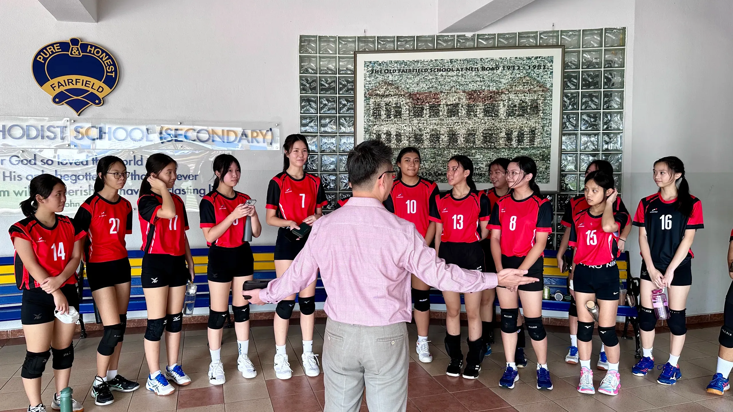 Members of the Fairfield girls' volleyball team stand in a wide semi-circle, listening intently as our principal, Mr Wee Tat Chuen in a pink long-sleeved shirt and trousers speaks with his arms outstretched. The students are dressed in red and black jerseys with matching shorts and knee pads. Behind them, the school crest reading "Pure & Honest Fairfield" and a framed historical artwork of the school building are visible on the wall.