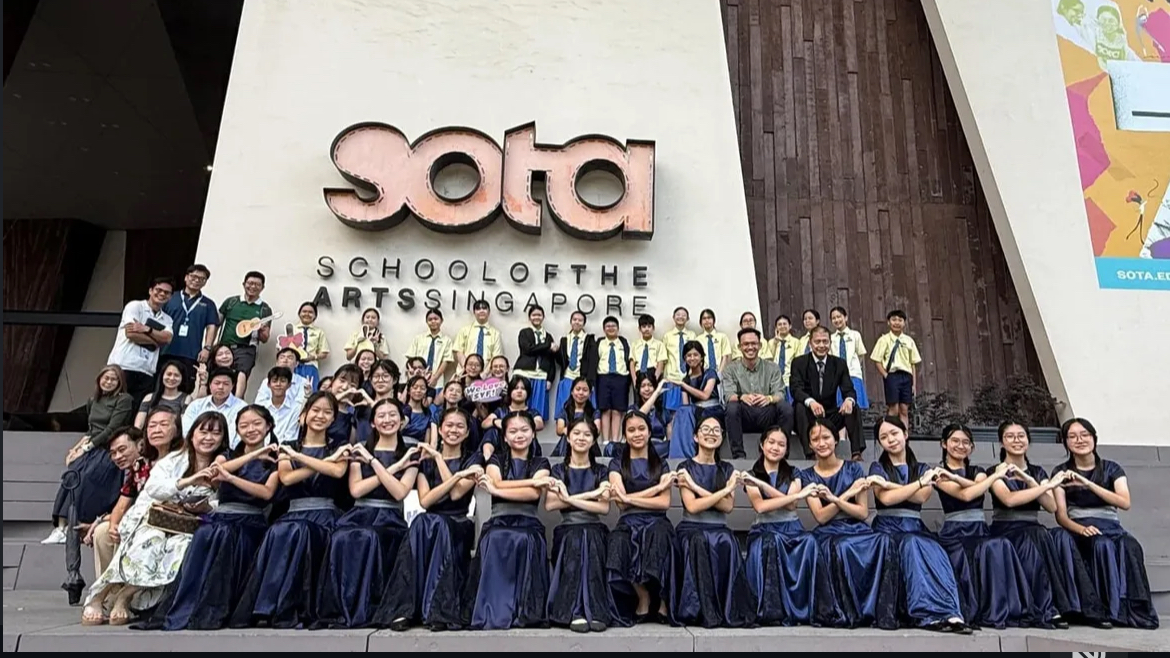 Members of the school guitar ensemble gather for a group photo on the concrete steps outside the School of the Arts (SOTA) Singapore during SYF. In the front row, female students dressed in shiny navy blue performance gowns sit cross-armed or making heart shapes with their hands. Behind them stands another row of students wearing light yellow school shirts with ties and blue skirts or trousers. On the right side of the middle row, the ensemble instructor sits smiling in a formal black suit next to another male teacher. On the far left, a group of teachers and parent supporters pose alongside the students. The prominent "SOTA" logo is affixed to the textured wall in the background.