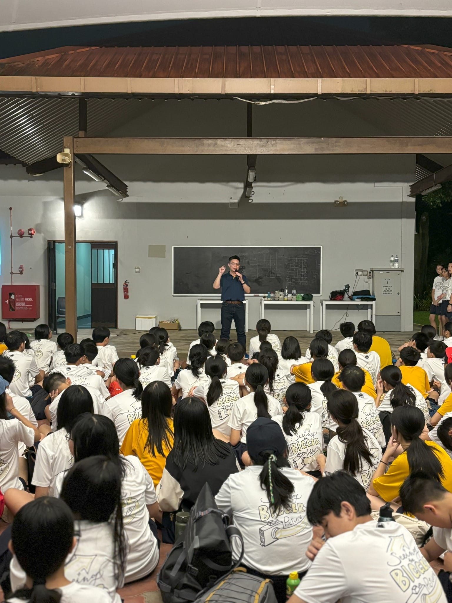 An evening camp session where school principal Mr. Wee Tat Chuen stands under a lit outdoor campsite pavilion, speaking into a microphone. He is dressed in a dark blue polo shirt and spectacles. A large cohort of Secondary One students are seated cross-legged on the floor in neat rows, listening attentively. Most students are wearing white camp t-shirts featuring a graphic on the back that reads "Seeing the Bigger Picture," while others are in yellow school sports shirts. The dark night sky and outdoor campsite surroundings are visible beyond the sheltered pavilion.