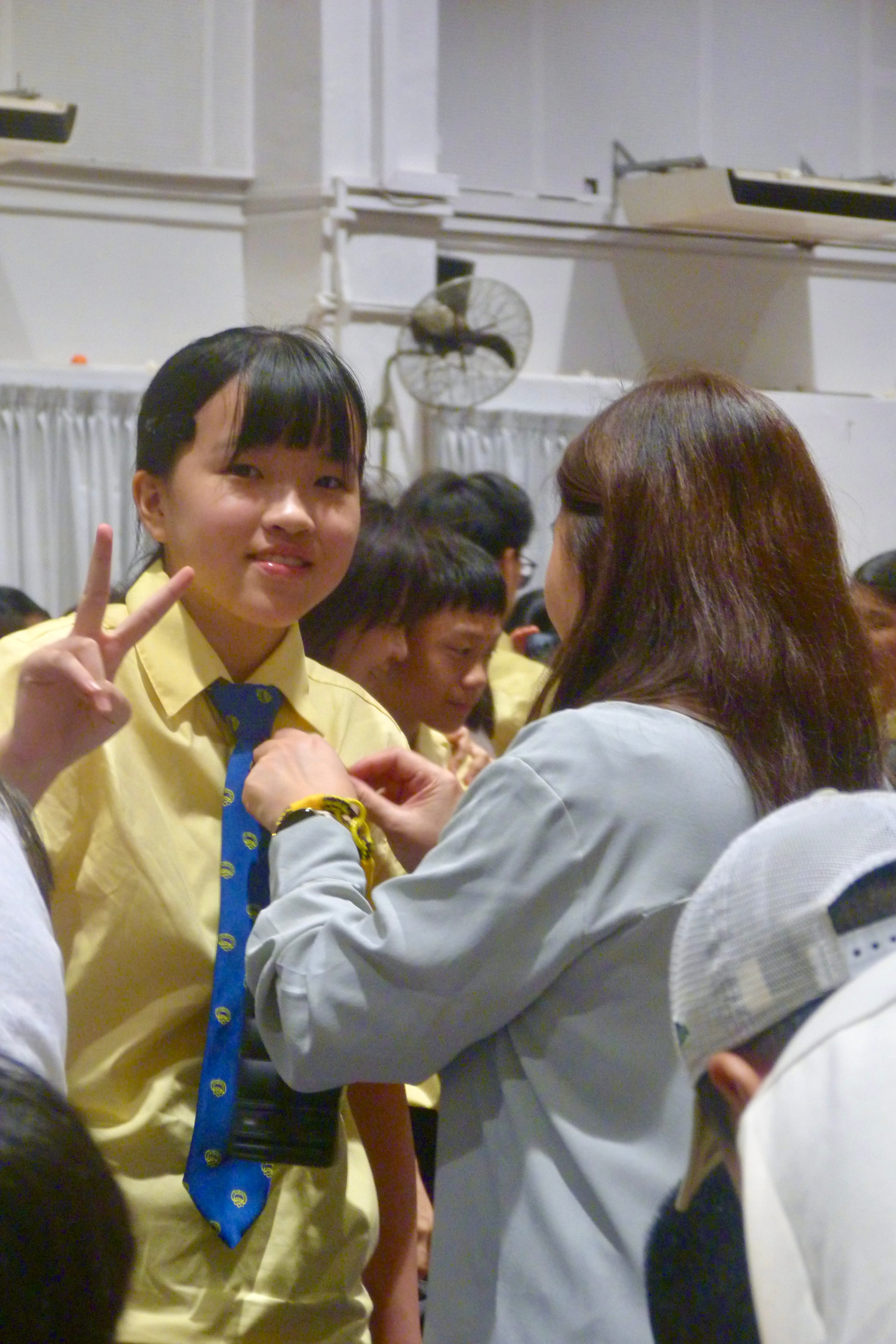 A female Secondary One student smiles and flashes a peace sign as her mother pins the school badge onto her uniform blouse during the traditional Installation Night ceremony in the school hall.