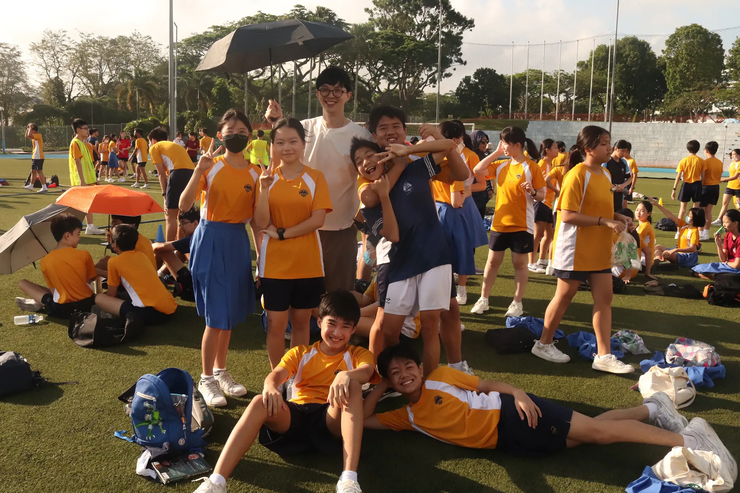 A group of Fairfield Methodist School (Secondary) students and a male teacher, Mr Kua posing together on a green synthetic field under sunny skies at Ngee Ann Polytechnic. The students are dressed in their yellow and white school PE uniforms, flashing peace signs and smiling warmly, while their classmates rest on the field with umbrellas in the background.