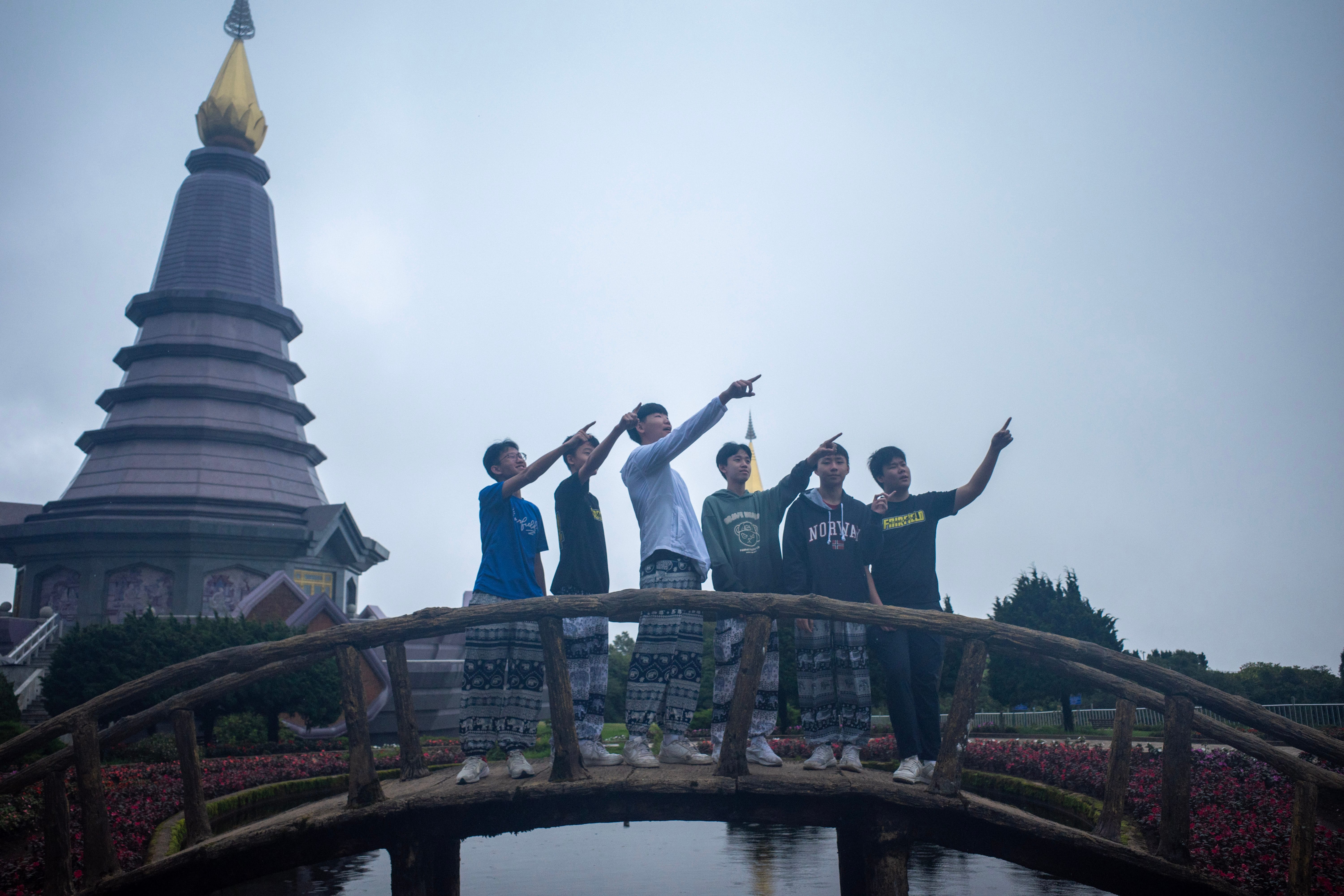 ix Secondary 3 students on a Trips for International Experience (TIE) journey pose together on a rustic, arched wooden footbridge over a small pond in Thailand. The students are lined up side-by-side, each smiling and pointing their right arm upward toward the sky in a synchronized gesture. Most of them are wearing loose, black-and-white patterned traditional Thai elephant pants paired with t-shirts and hoodies. In the background, a large, multi-tiered dark grey pagoda with a prominent gold spire rises against a misty, overcast sky, surrounded by landscaped flowerbeds.