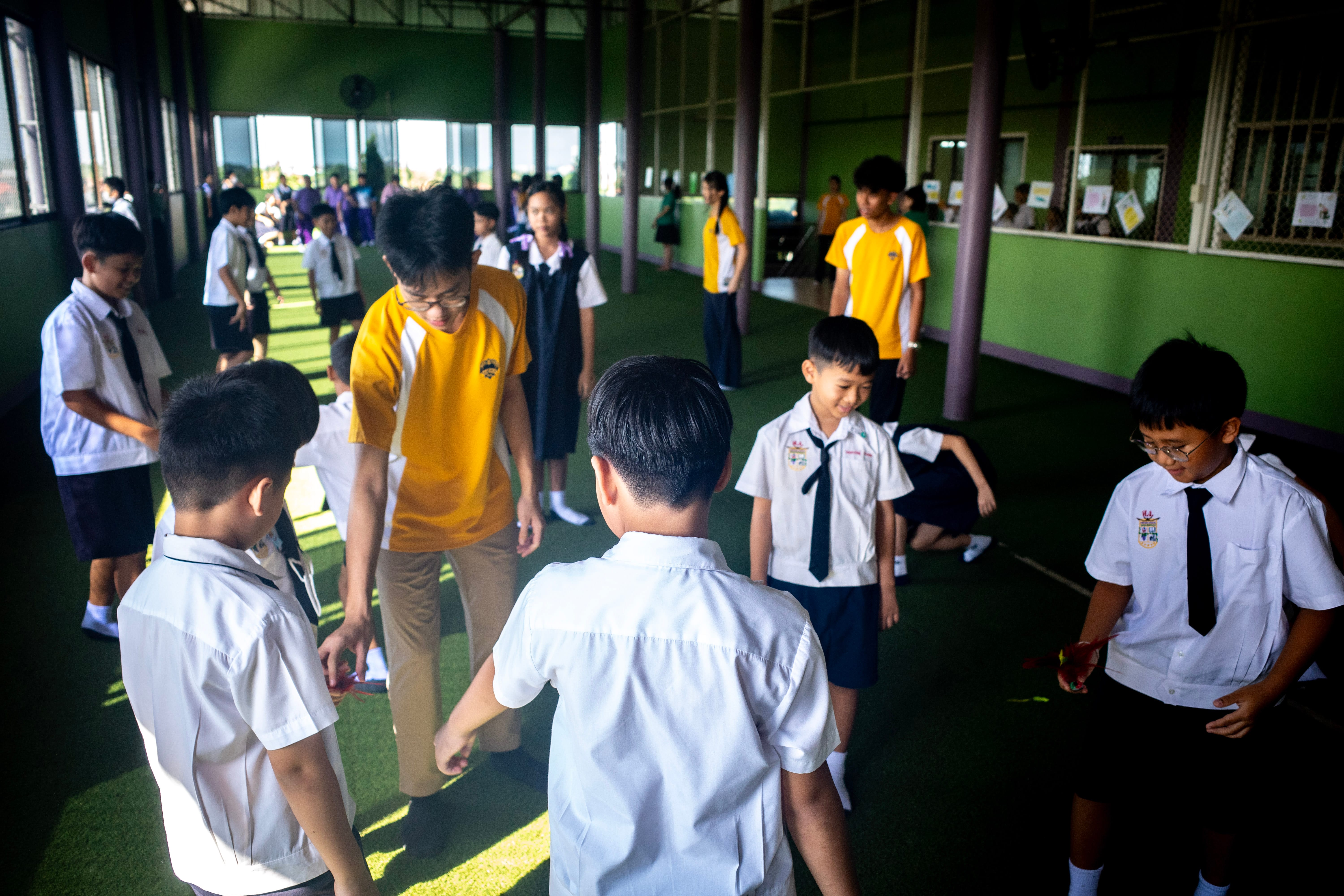 A group of Secondary 3 students interact with local primary school children in a large, green-carpeted indoor activity hall during their TIE learning journey in Thailand. In the foreground, a male secondary student wearing a yellow and white school sports t-shirt and khaki pants leans down to hands-on guide a group of younger Thai boys dressed in traditional white school shirts and dark shorts. Other groups of primary school students are scattered across the spacious hall, playing icebreaker games together near the green walls and support pillars while sunlight streams through the wide side windows.