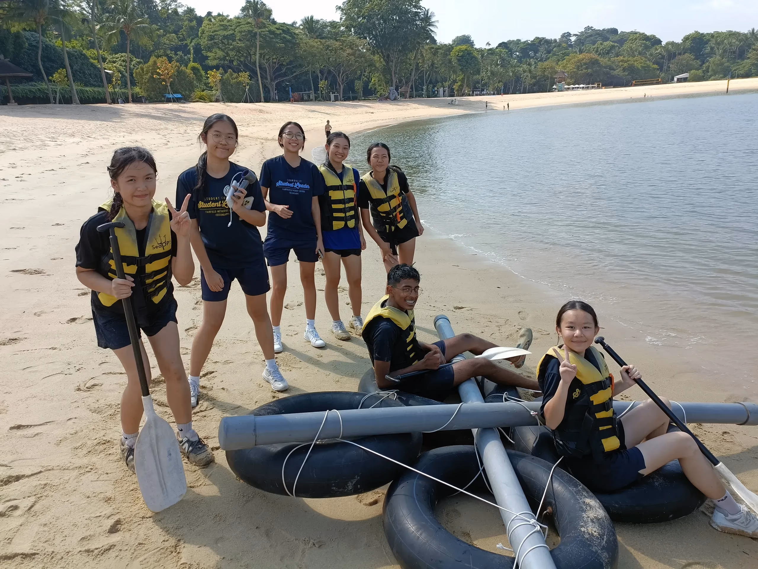 Seven student leaders from Fairfield Methodist School (Secondary) posing together with a self-built raft on a sandy beach during the Sophia Blackmore Leadership Programme training camp in 2025. Two students are positioned on the raft—a boy sitting near the center and a girl on the right giving a thumbs-up while holding a paddle. Five girls stand behind them on the shore, some wearing life jackets, smiling and making peace signs. The raft is constructed from large black inner tubes lashed to grey PVC pipes. In the background, calm ocean water meets a curved sandy coastline lined with lush green tropical trees.
