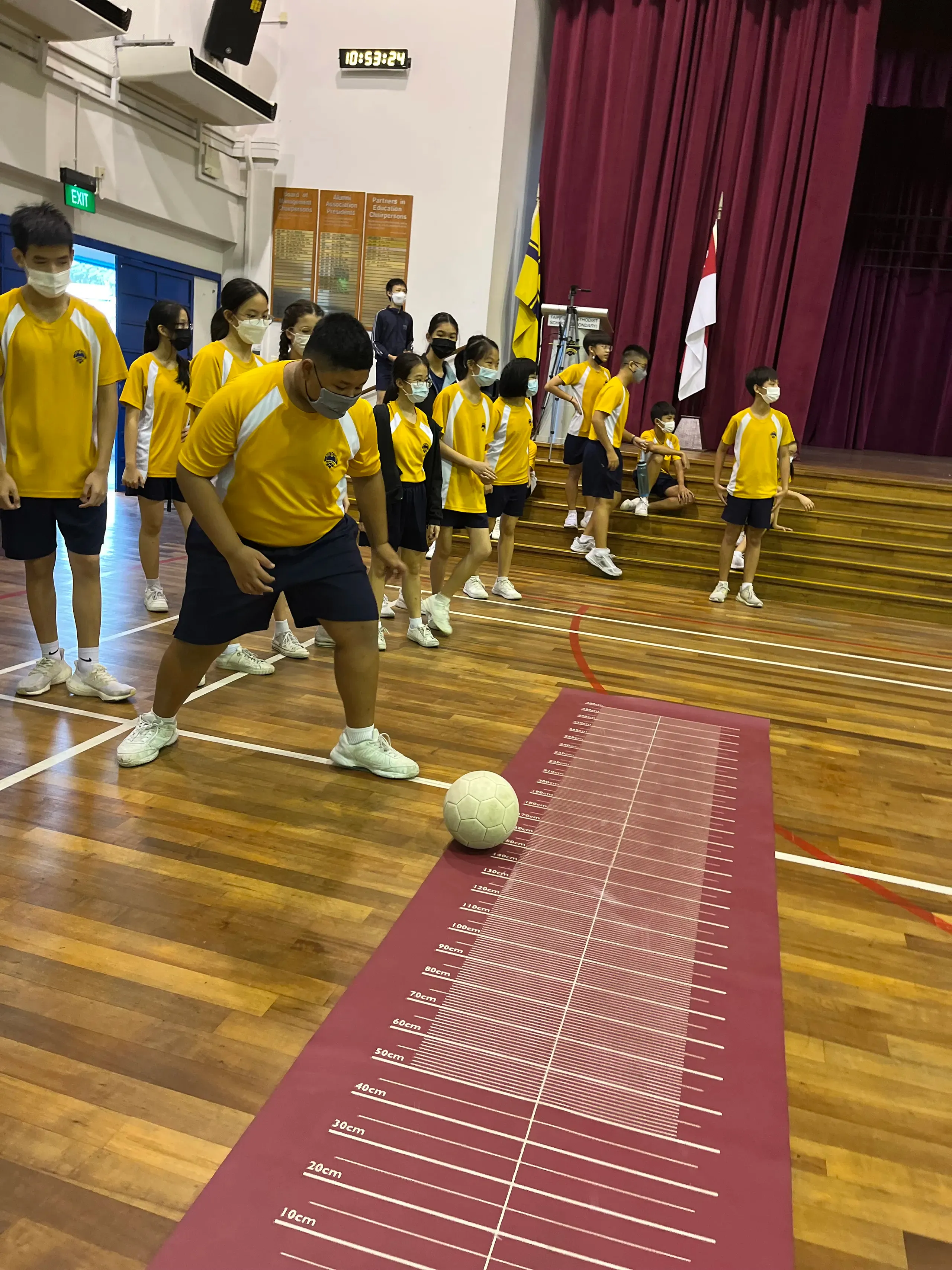 Inside a school hall during FLEX week, a masked male student in a yellow and blue sports kit prepares to roll or kick a football down a long, red measurement mat rolled out on the wooden floor. His classmates stand in line behind him or sit on the steps of the stage in the background, watching the activity. The school stage features a dark red curtain and flags on display.