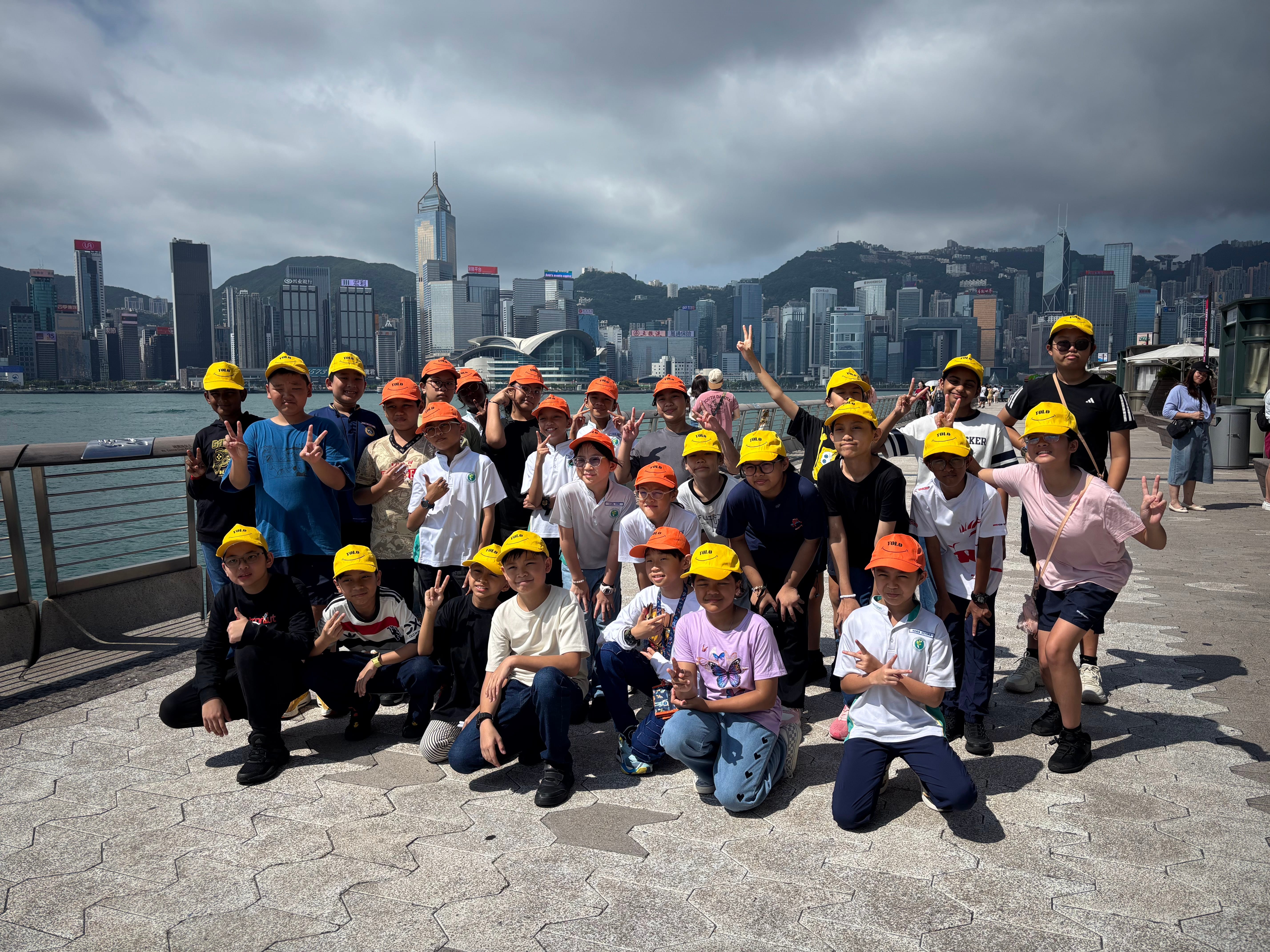 Group of kids in orange and yellow caps posing by the waterfront with a city skyline under dark clouds.