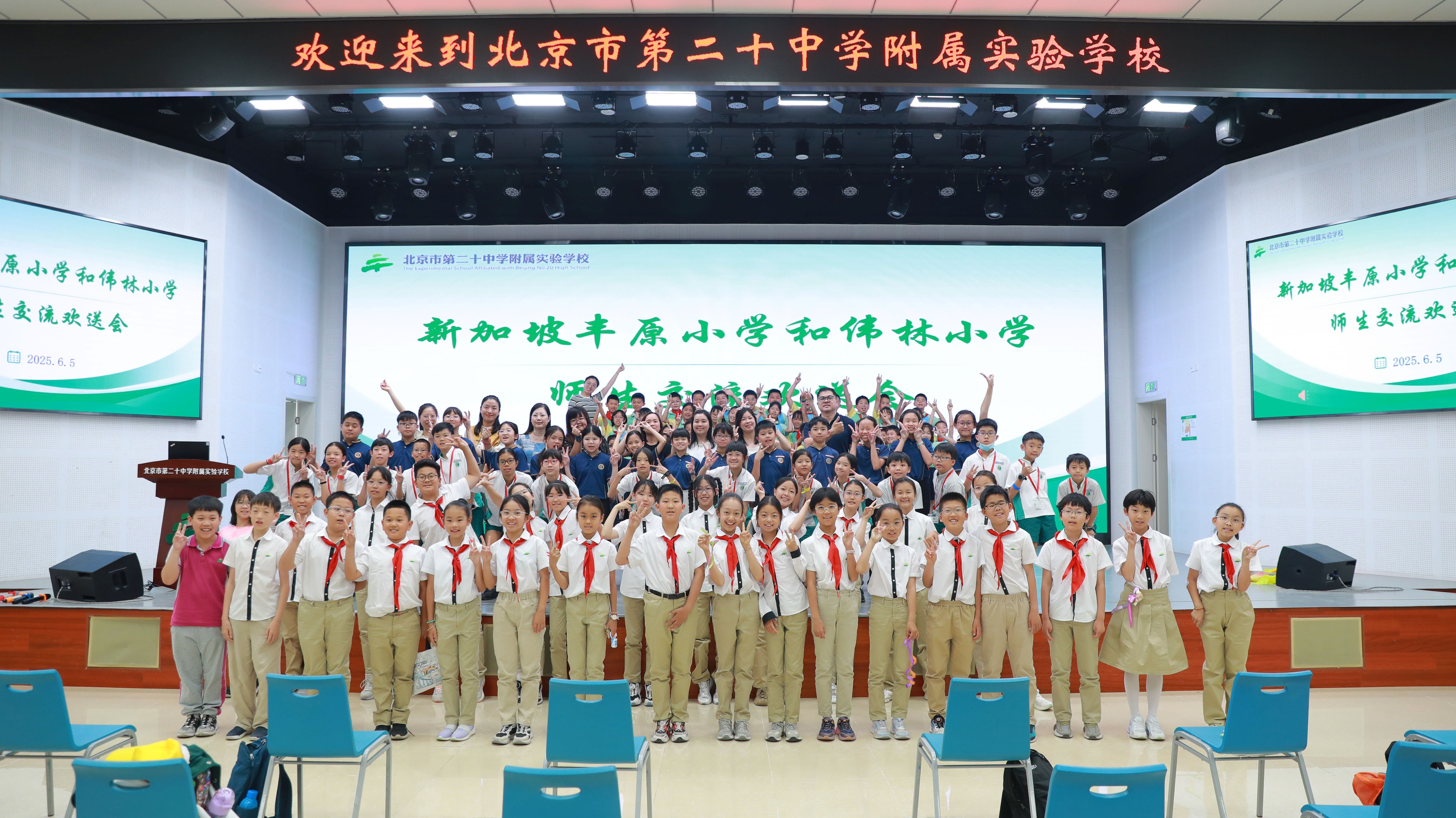 A large group of students and teachers pose cheerfully in a Chinese school auditorium with Chinese text on screens behind them.