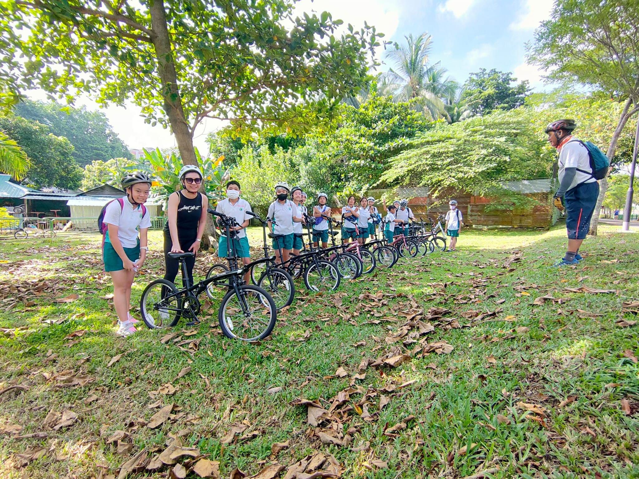 A group of children in helmets stand with bicycles lined up on grass in a park, listening to an instructor.