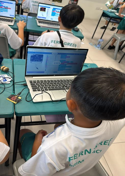 Children in white uniforms sit at teal desks using laptops with block-based coding software in a classroom.