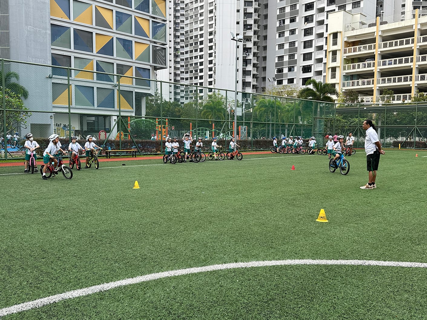 Young children in helmets line up on bicycles on a green turf field while an adult instructor watches nearby.