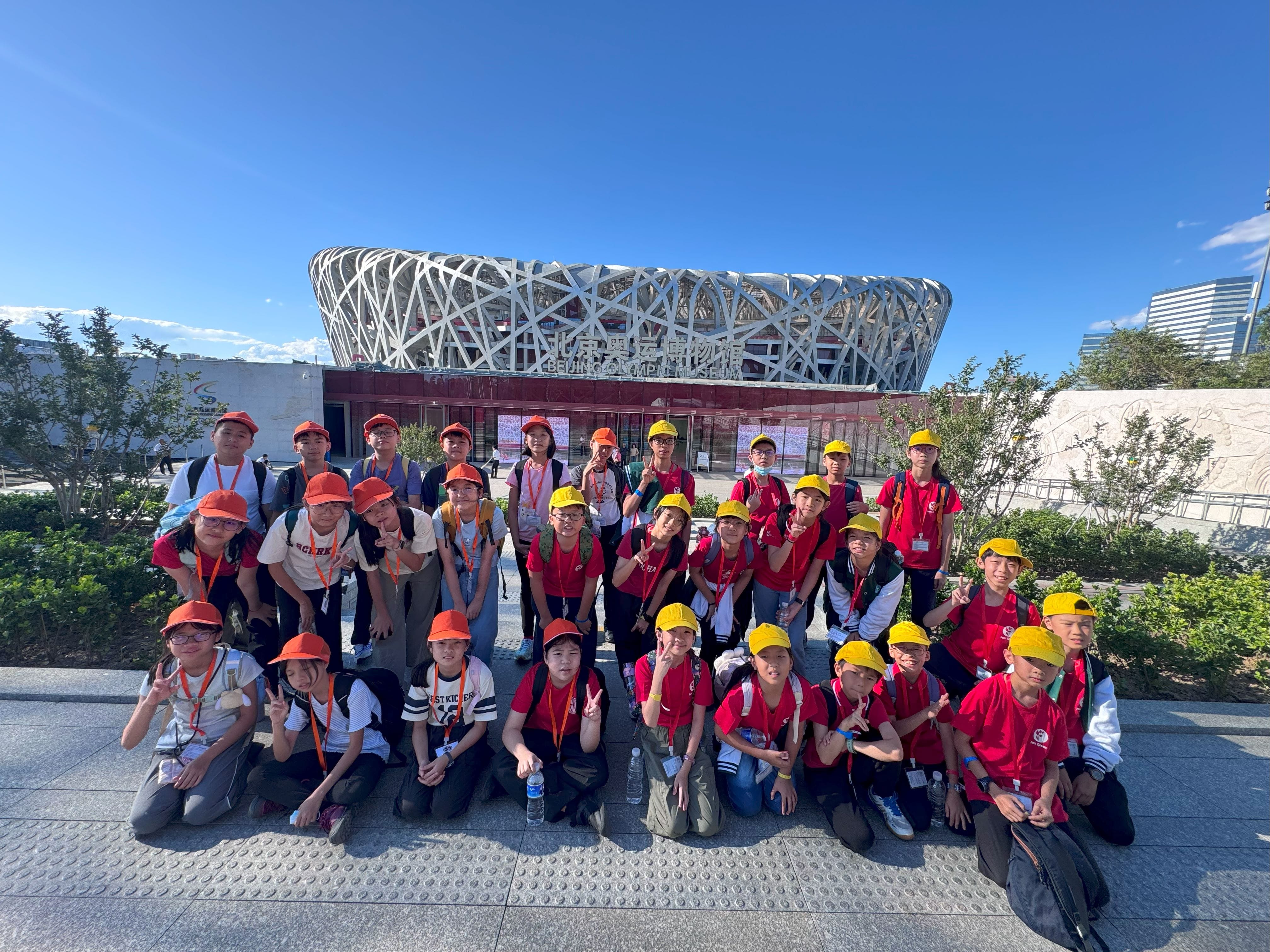 A group of children in red shirts and orange or yellow caps pose in front of the Beijing Olympic Museum and Bird's Nest stadium.