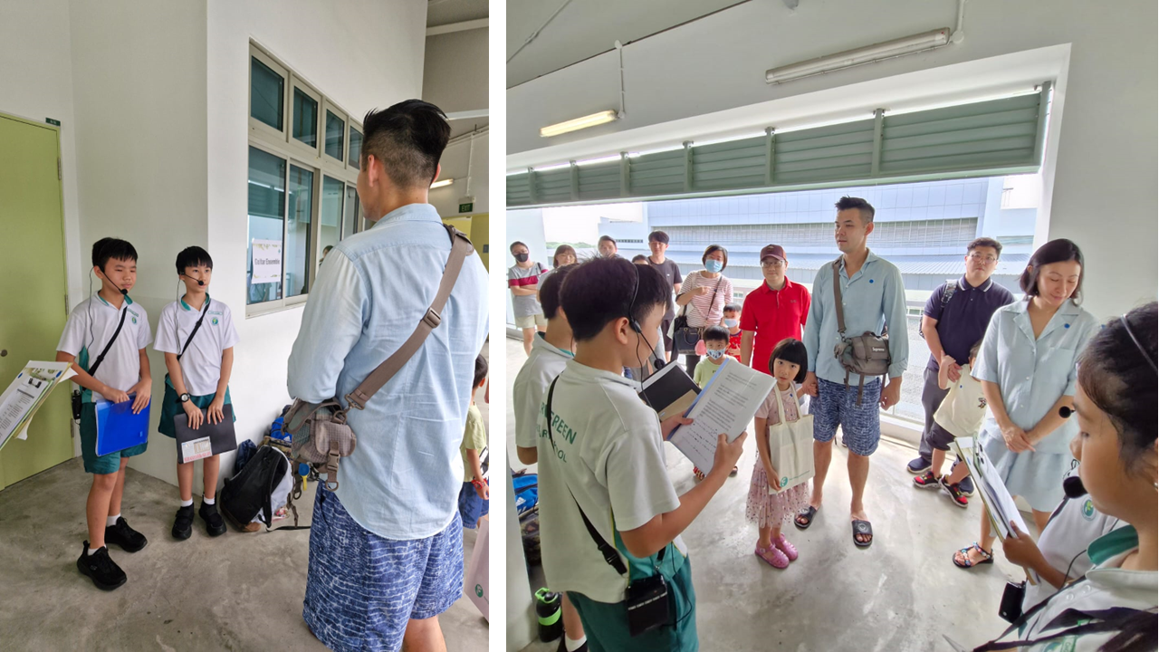 Students in school uniforms with microphones present to a group of adults and children indoors.