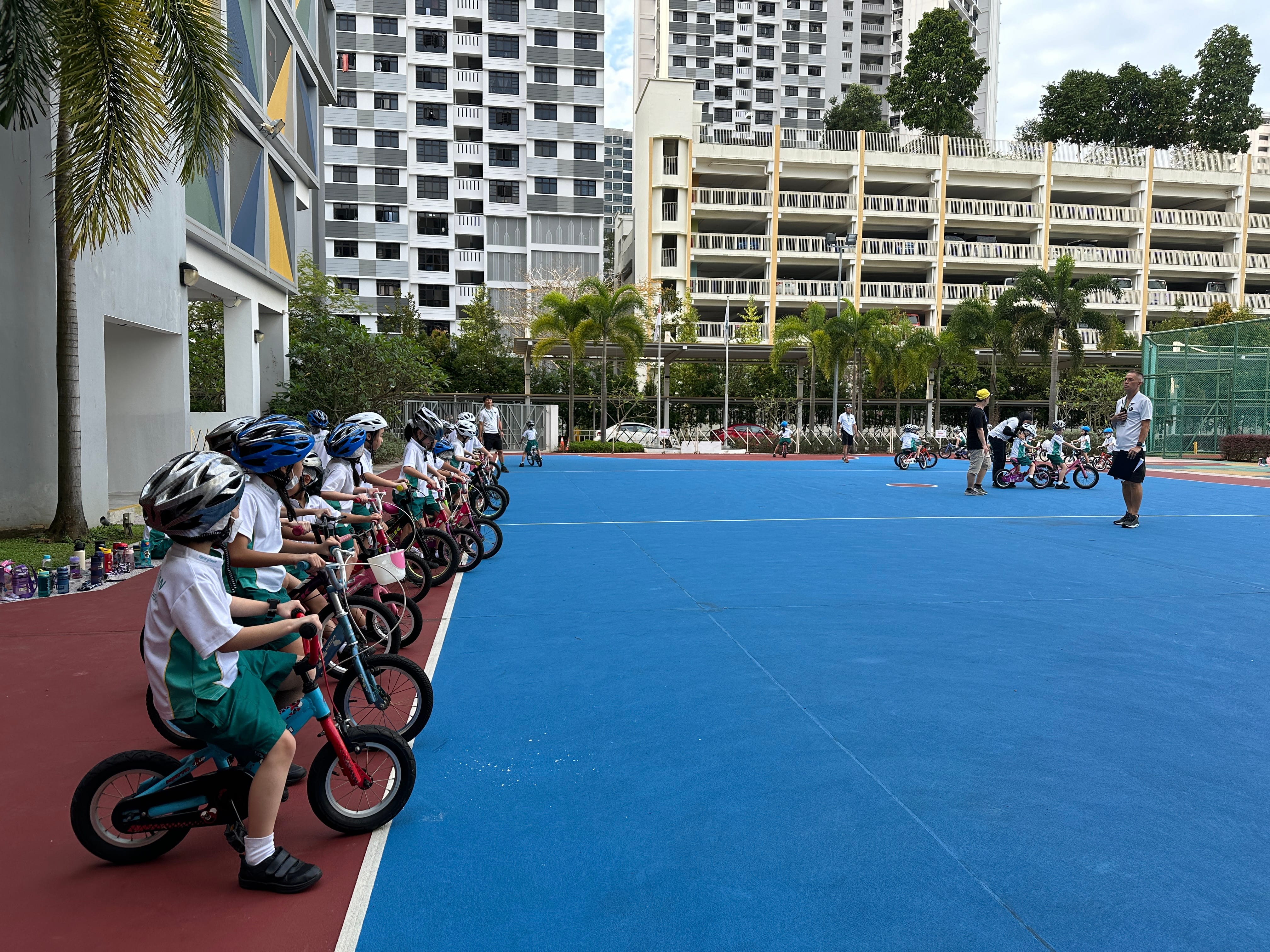 Children in helmets line up on small bicycles beside a blue court as two adults supervise in the distance