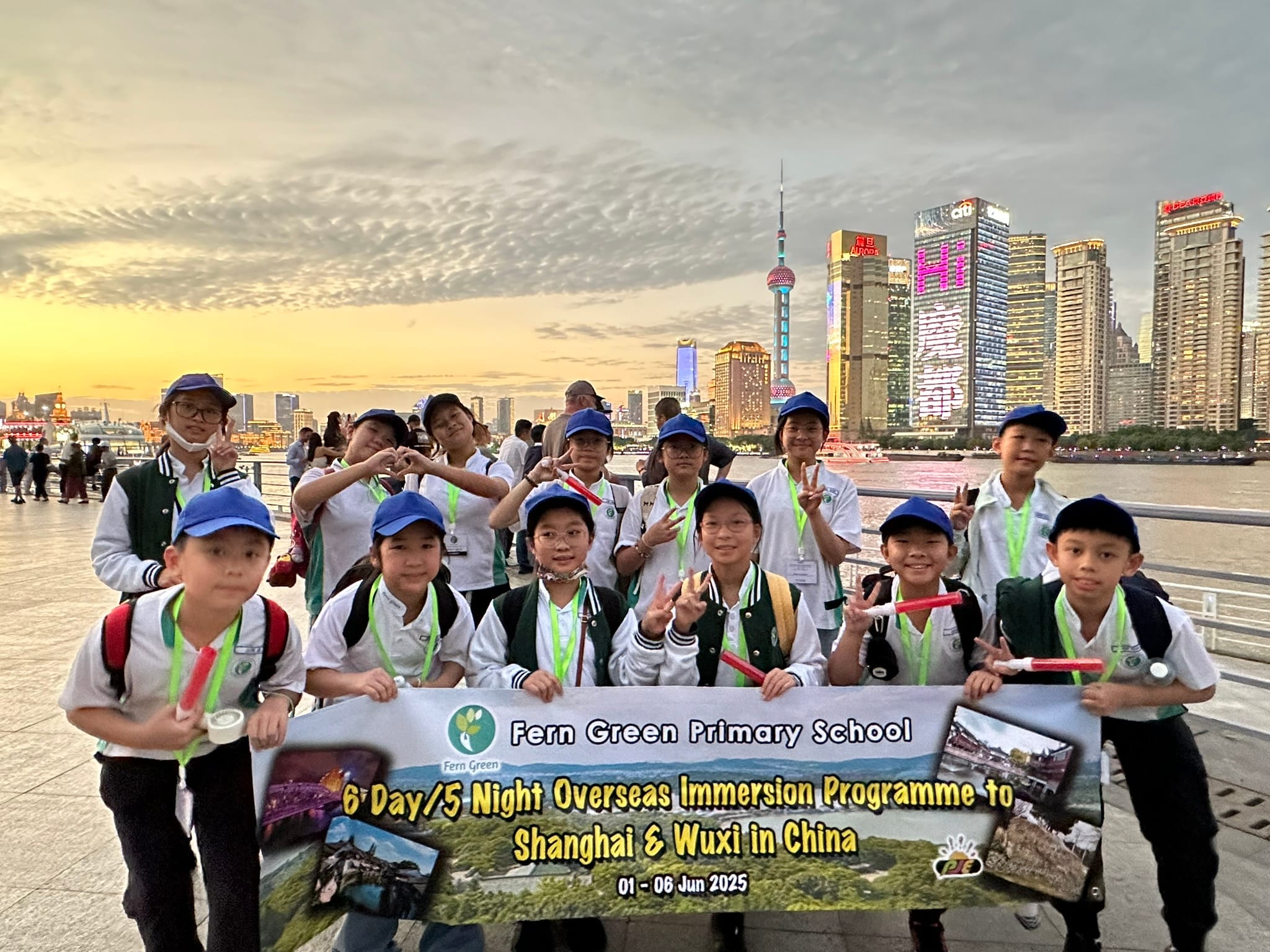 Students in blue caps hold a Fern Green Primary School banner on Shanghai's Bund with the lit skyline at dusk.