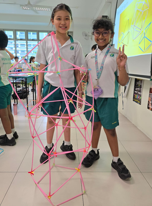 Two smiling schoolgirls in uniforms stand beside a tall pink straw geometric structure they built in a classroom.