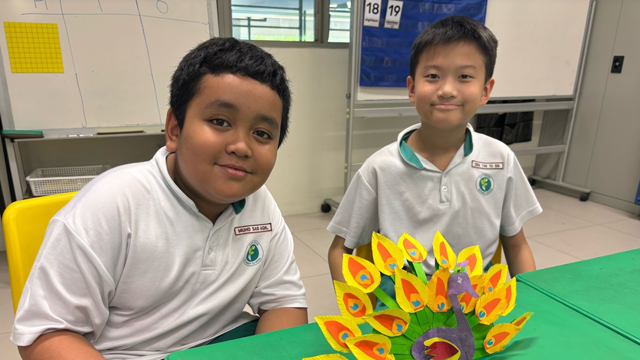 Two smiling schoolboys in white uniforms sit at a green table with a colorful paper peacock craft.