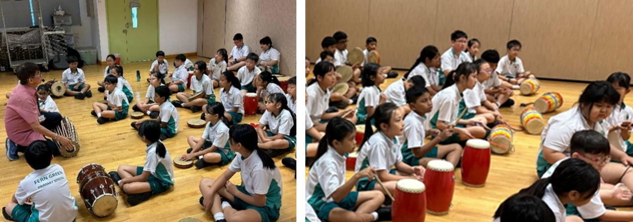 Students in white and green uniforms sit on a wooden floor with hand drums during a music lesson led by an adult.