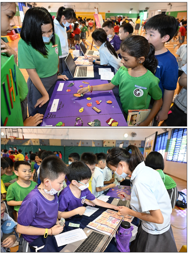 Children in green and purple uniforms gather around tables with learning activity boards and laptops in a school gymnasium.